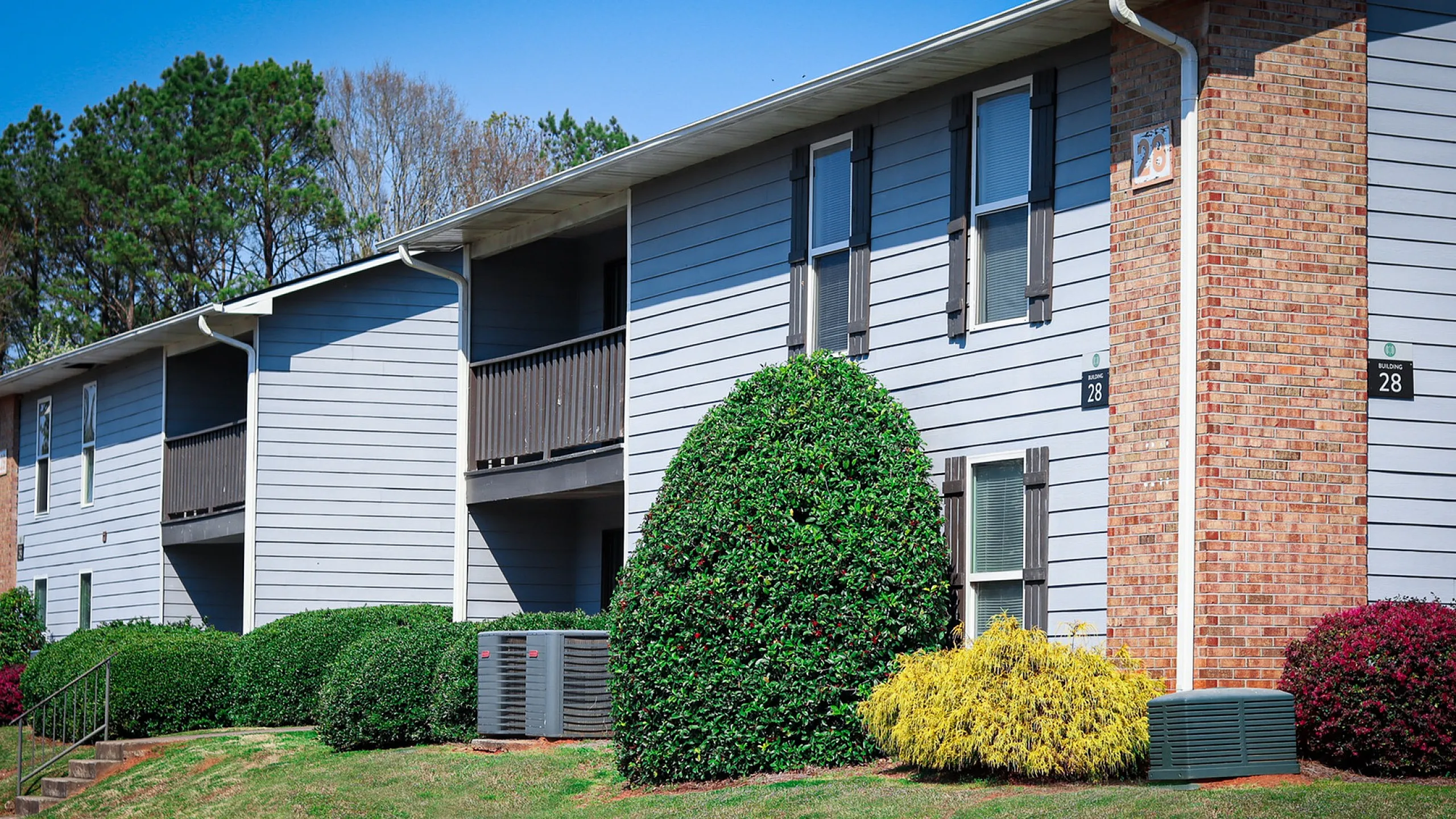 Charming apartment building with brick and siding exterior, featuring lush landscaping and private balconies, in a serene community setting.