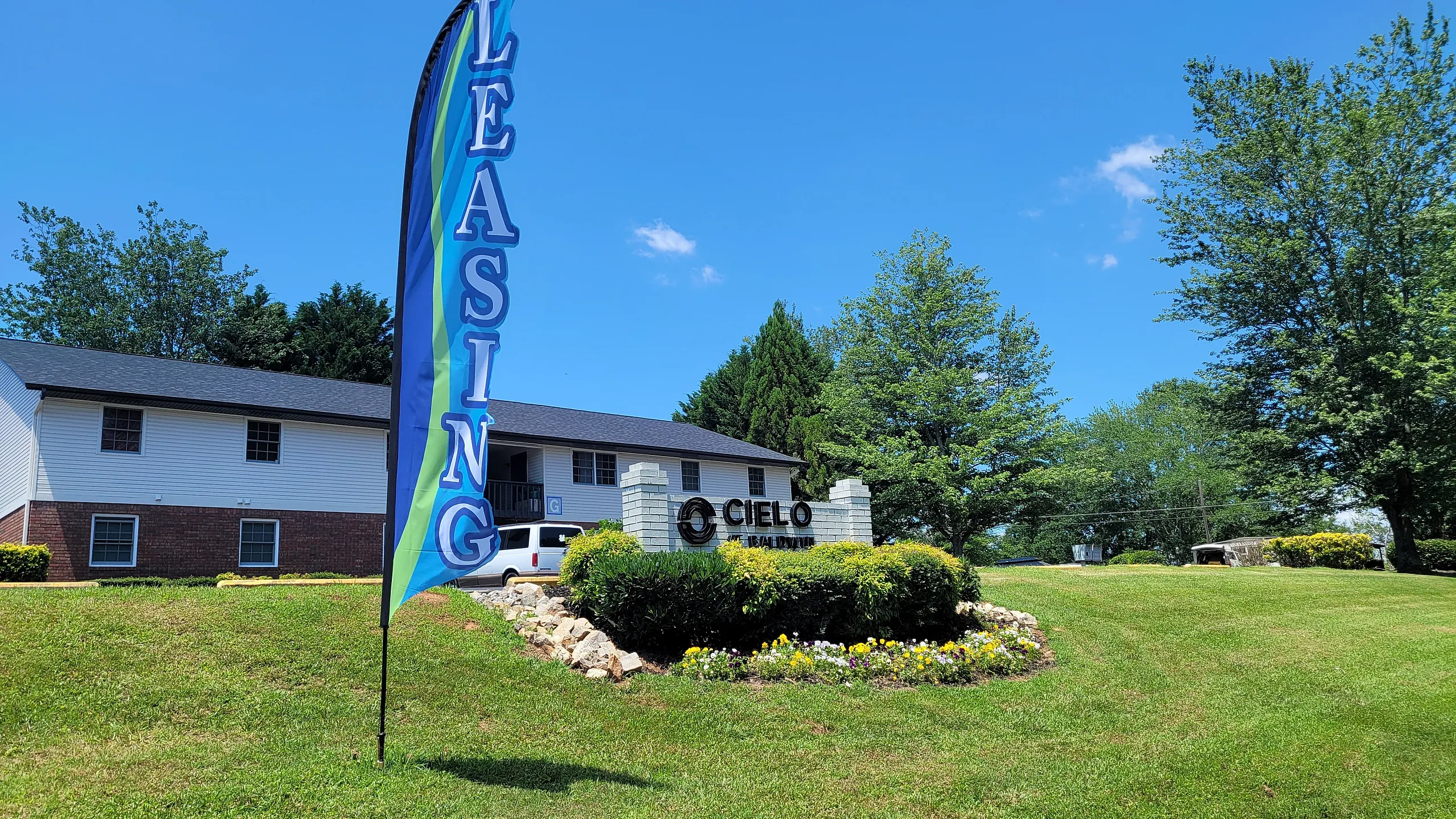 Charming apartment complex with vibrant 'Leasing' banner, surrounded by lush greenery and a sunny blue sky.