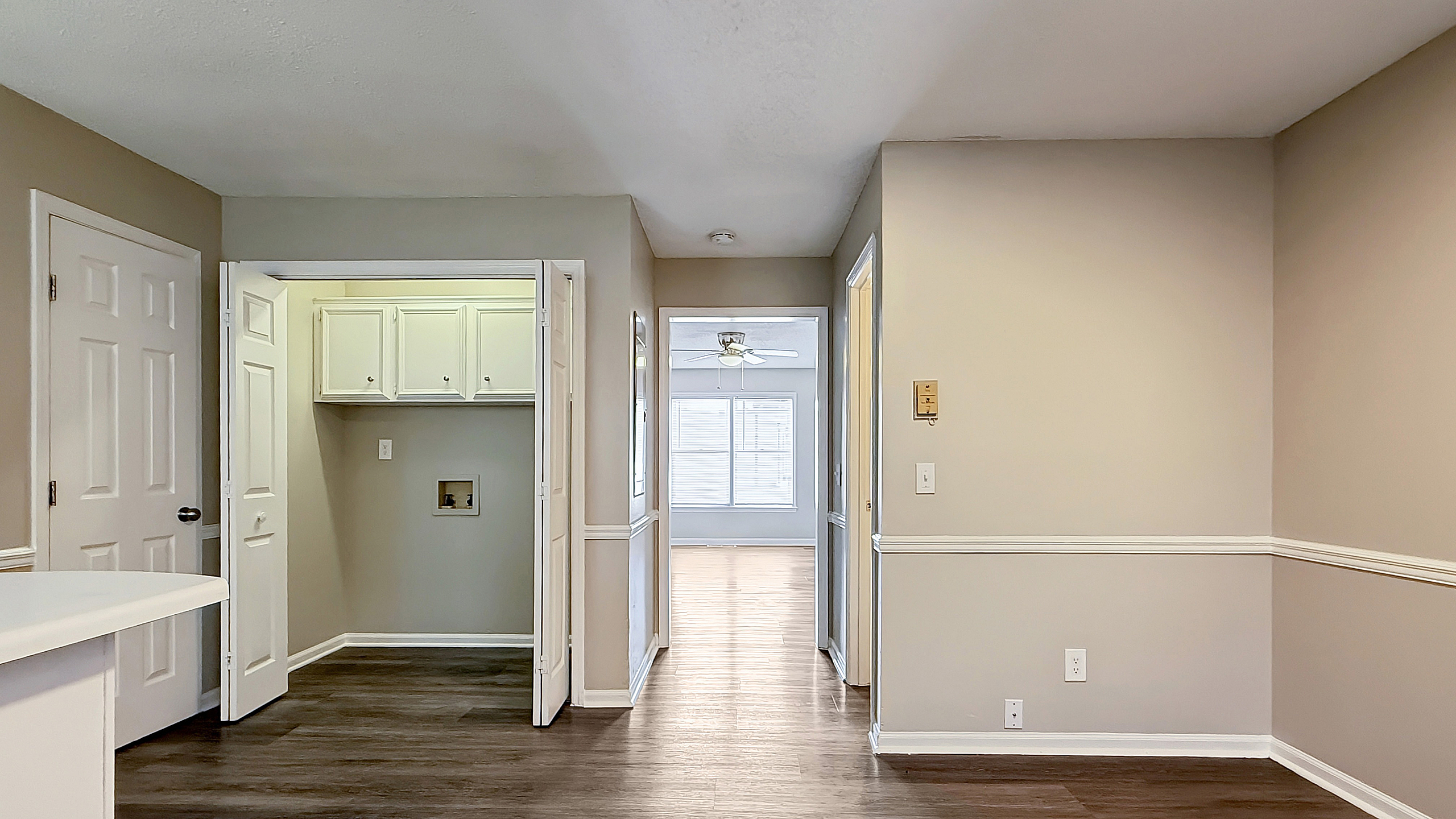 Bright, welcoming hallway with elegant wainscoting, showcasing a laundry area and leading to a sunlit room with modern flooring and ceiling fan.