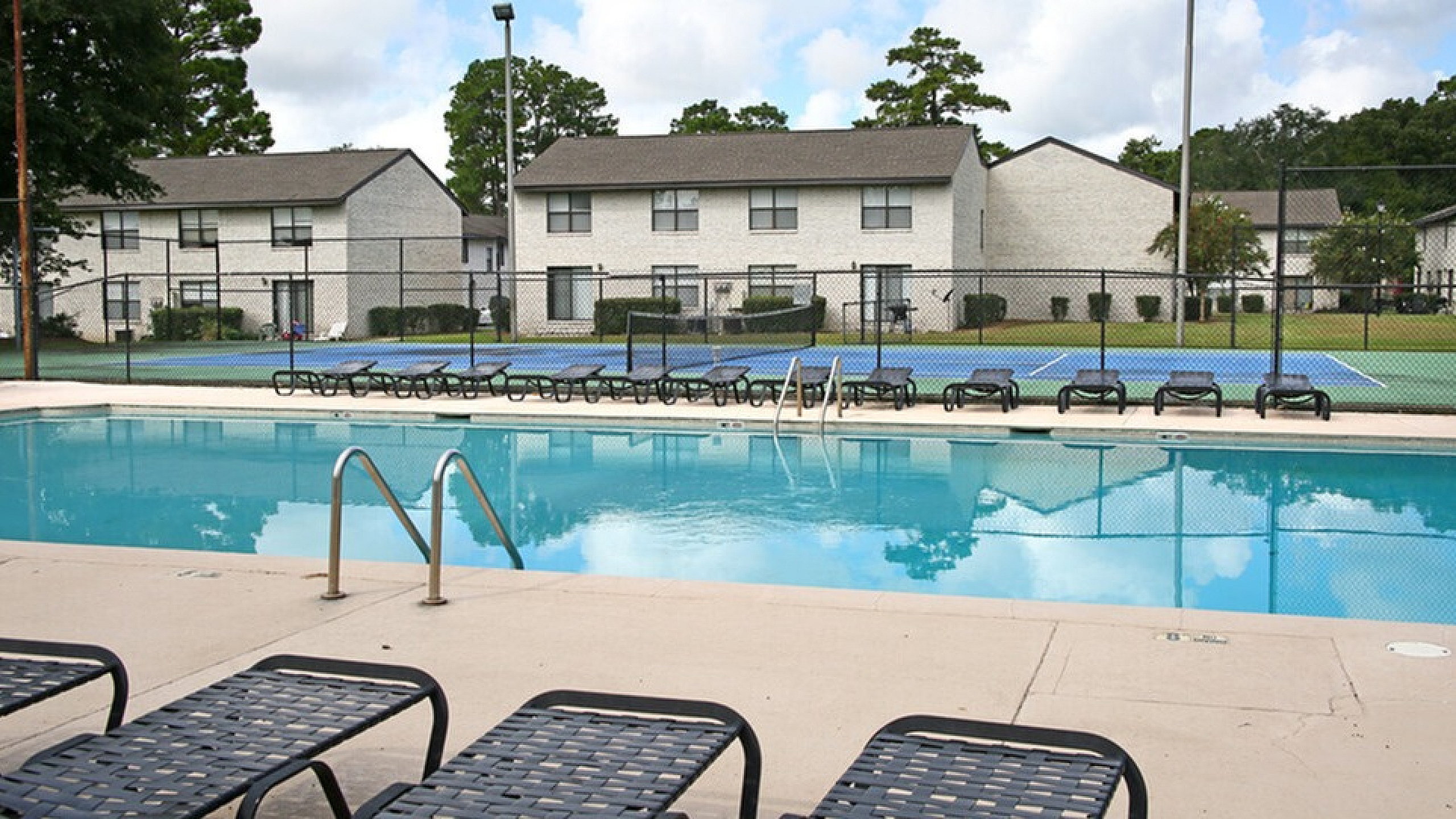 Inviting apartment complex featuring a refreshing pool, surrounded by stylish loungers and adjacent tennis courts with well-maintained landscaping.