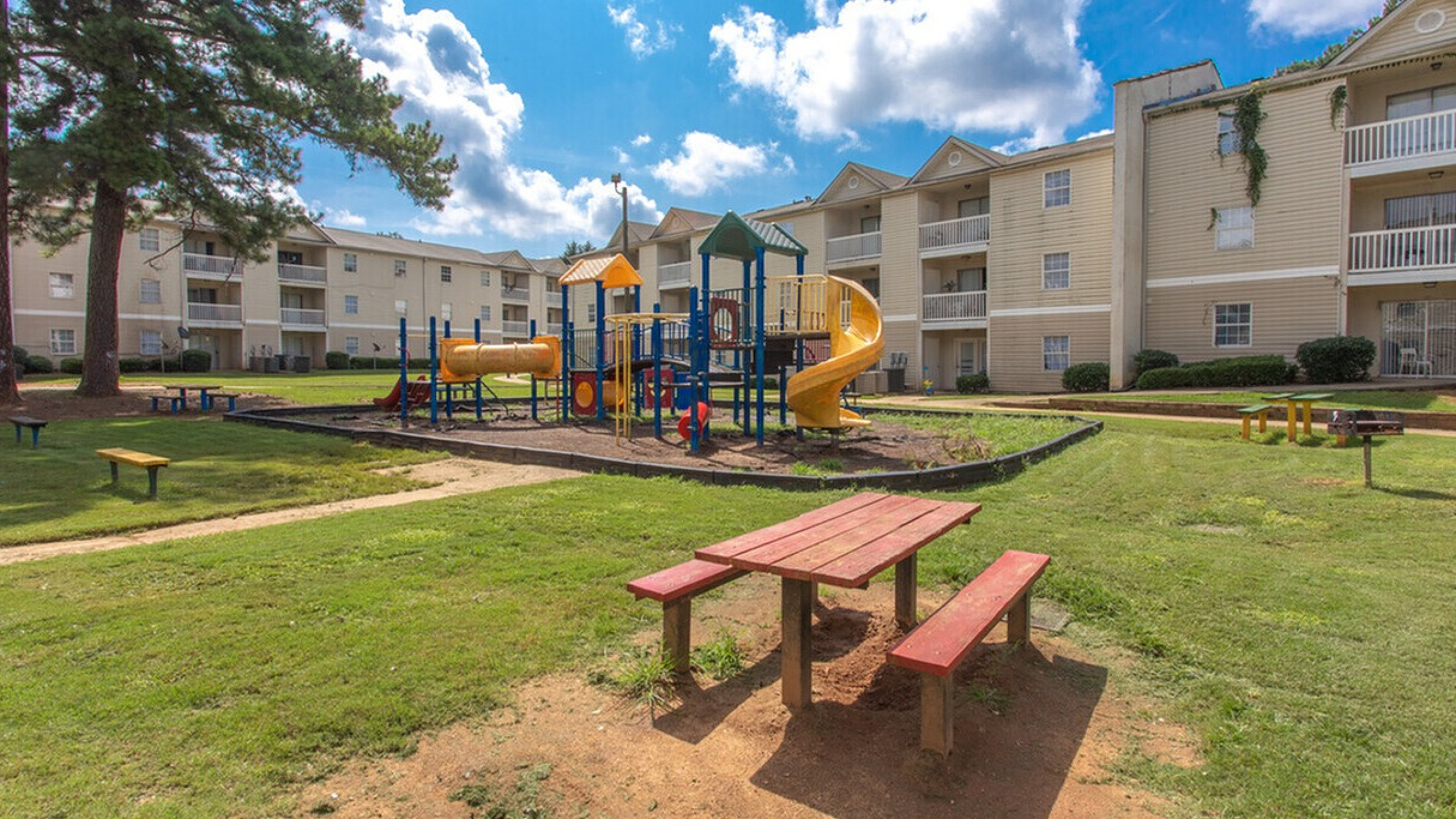 Inviting apartment complex with vibrant playground and picnic area, perfect for family enjoyment under sunny skies.