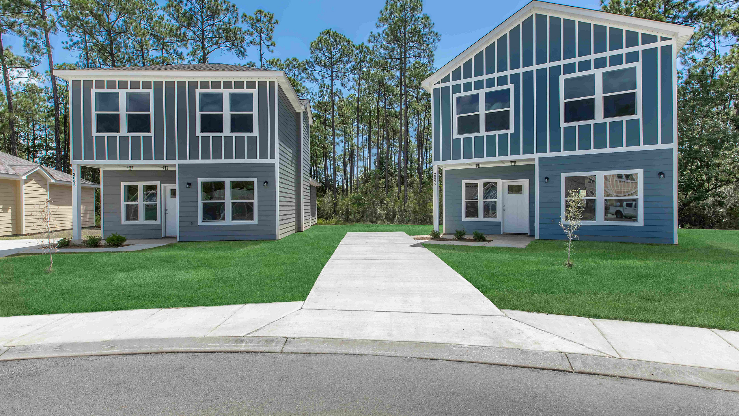 Charming modern duplex with vibrant blue siding, lush green lawns, and surrounded by serene pine trees under a clear blue sky.