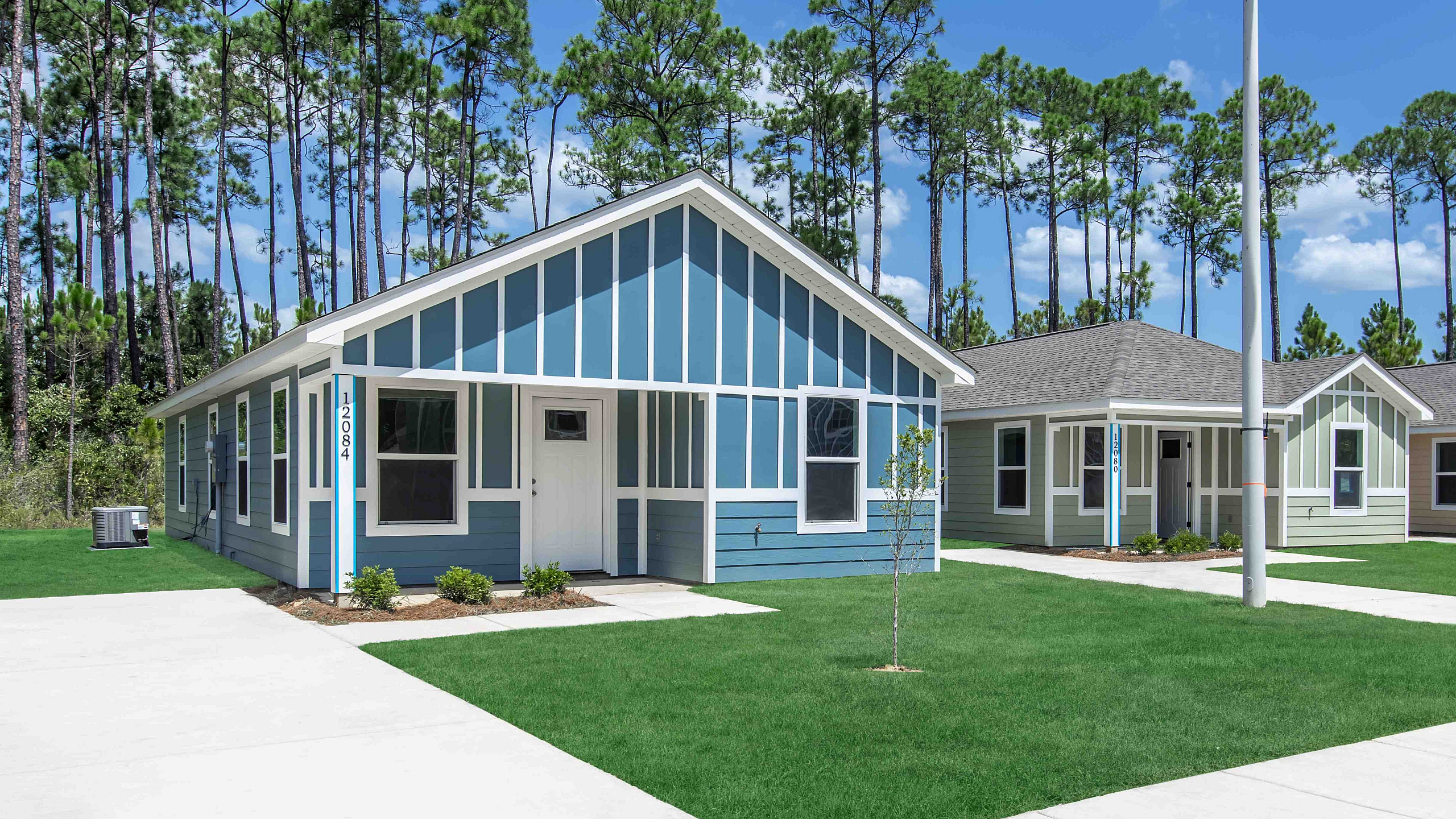 Charming blue cottage-style homes with vibrant lawns set against a backdrop of tall pine trees, offering a serene and picturesque neighborhood setting.