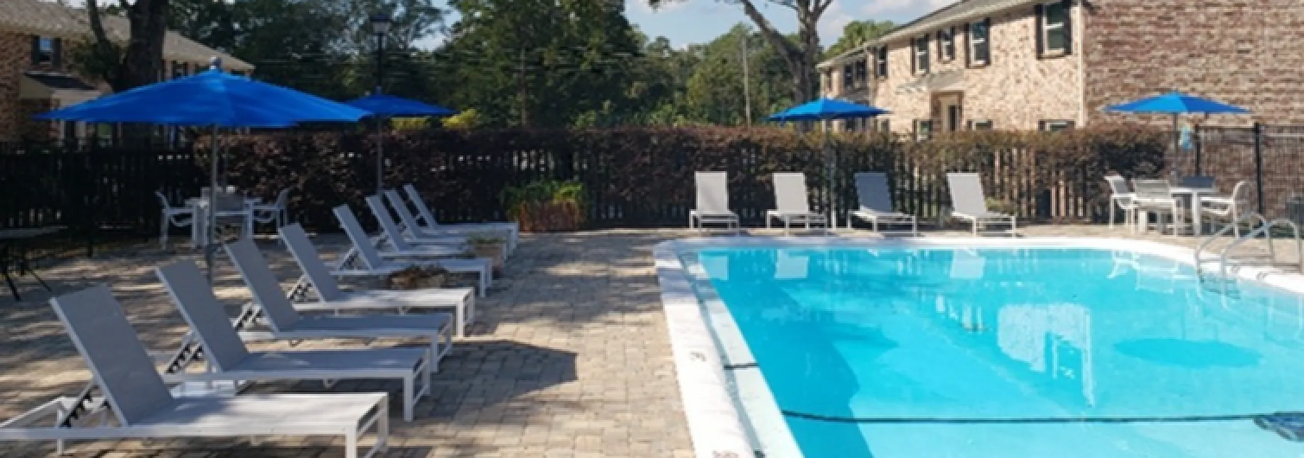 Inviting outdoor pool area with stylish lounge chairs and vibrant blue umbrellas, surrounded by charming brick buildings and lush greenery.