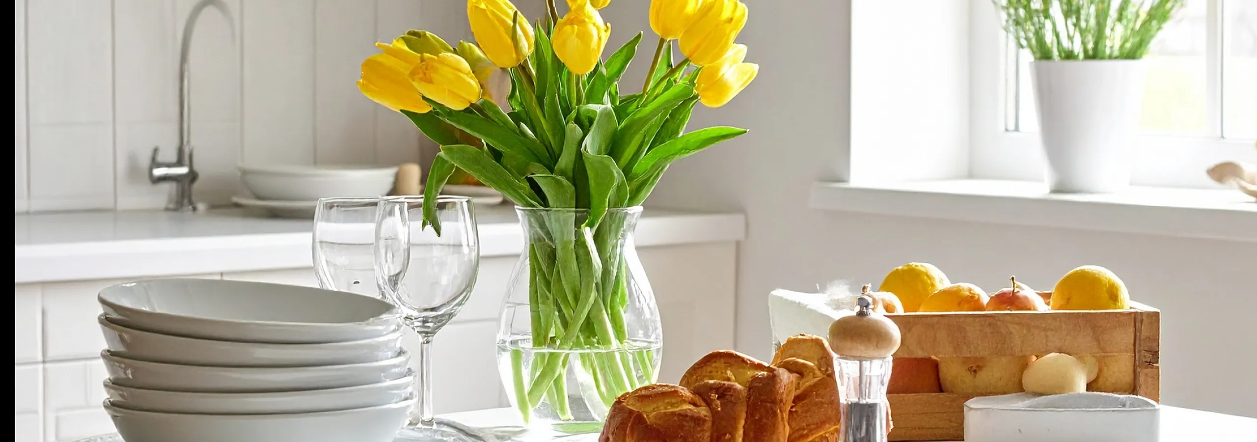 Charming white kitchen with bright natural light, featuring yellow tulips in a vase, elegant white dishware, fresh fruit basket, and artisanal bread, creating a warm and inviting atmosphere.