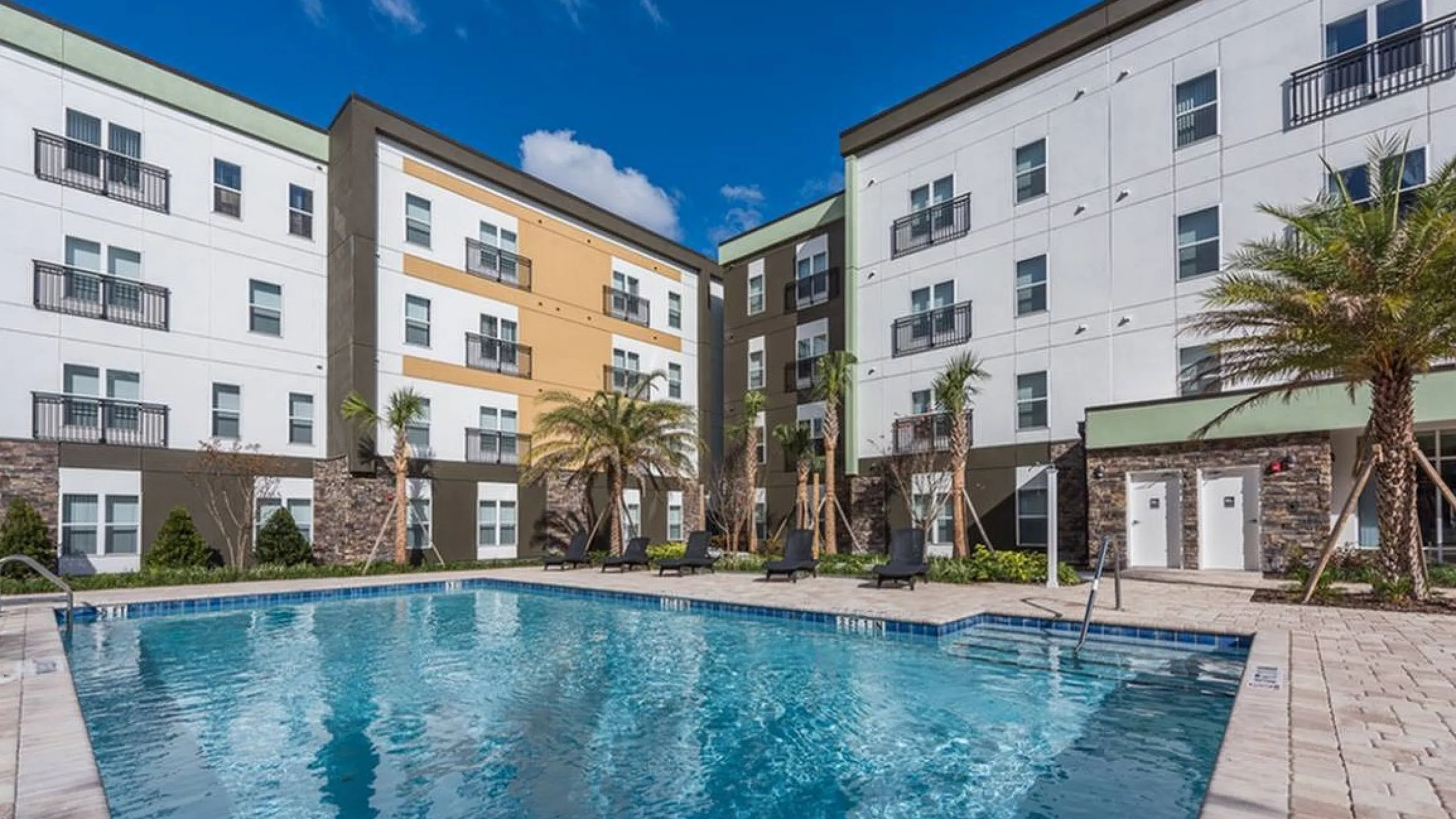 Modern apartment complex with a sparkling pool, surrounded by palm trees and stylish architecture under a clear blue sky.