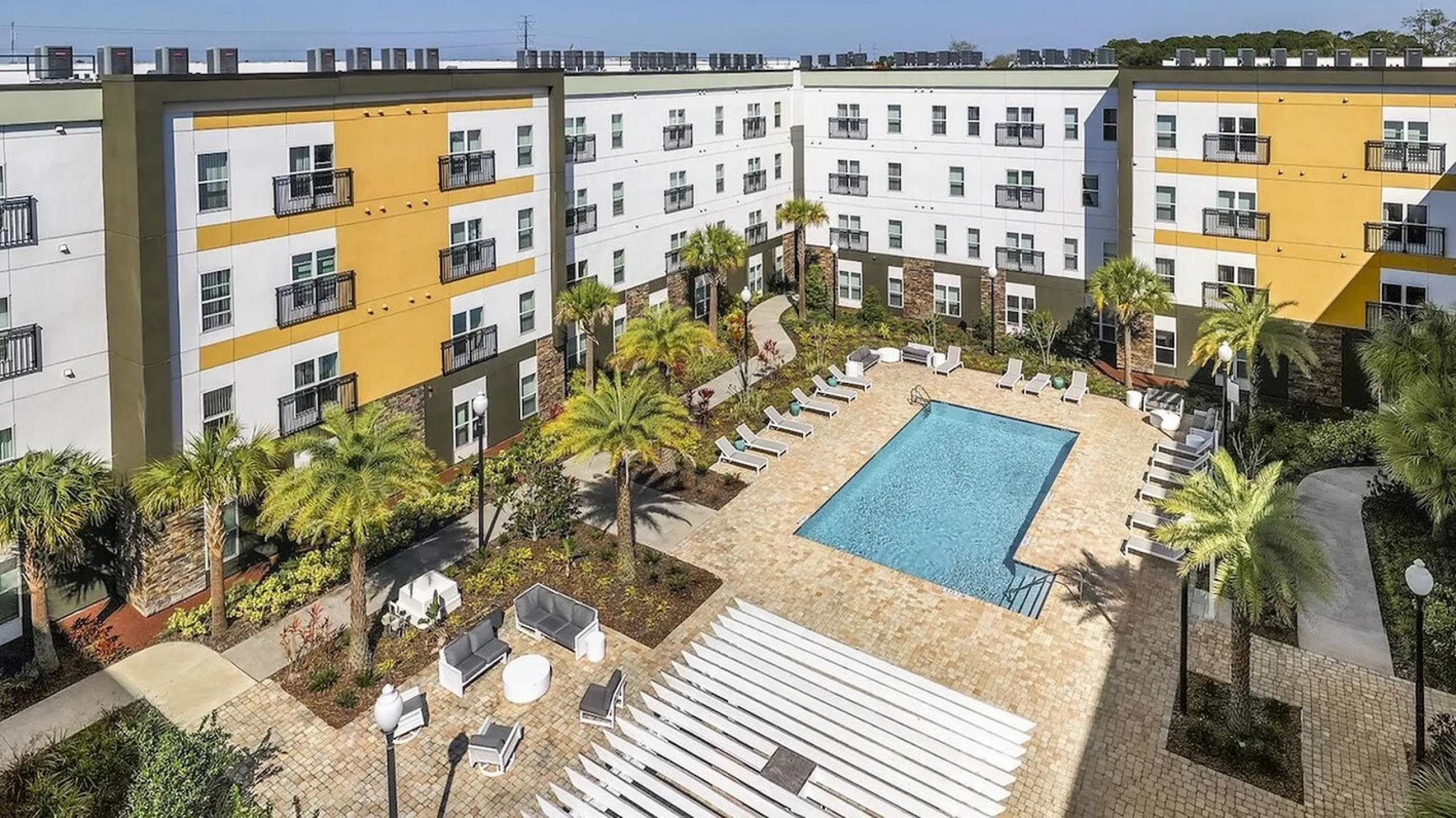 Stylish apartment complex featuring a central swimming pool surrounded by lush palm trees, modern buildings, and inviting lounge seating.