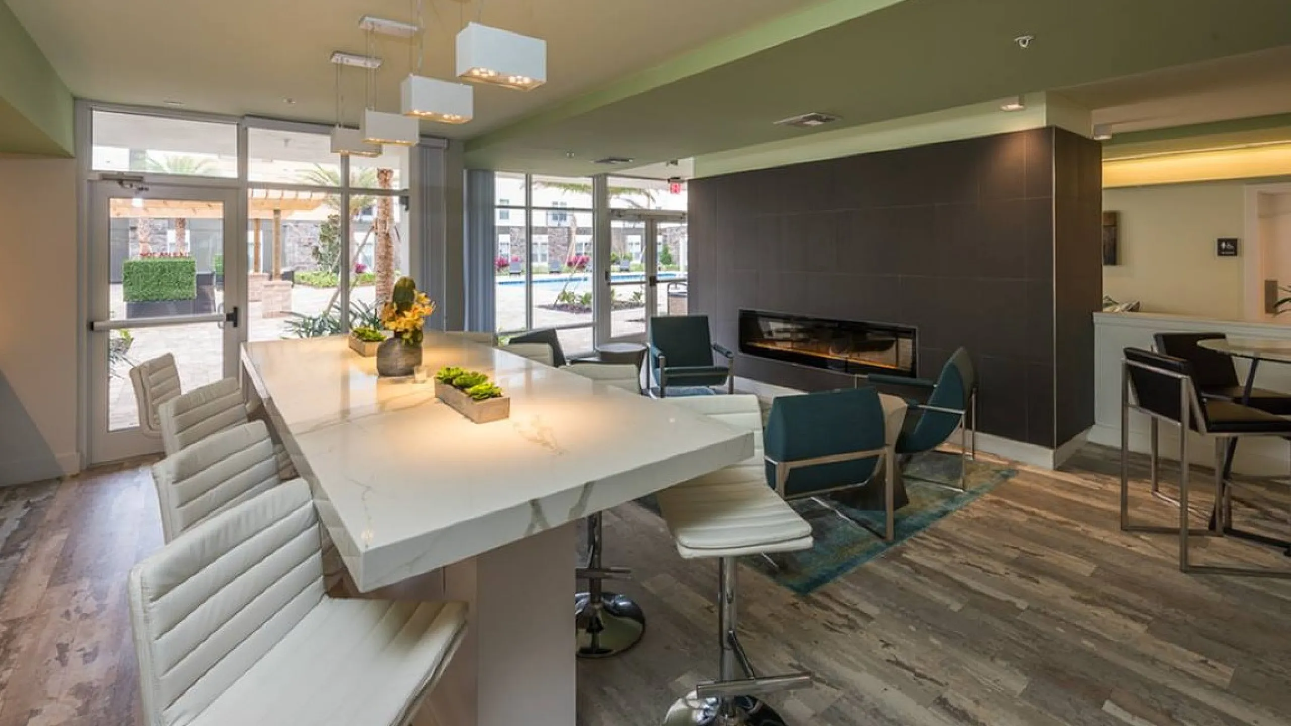 Modern apartment interior showcasing a luxurious kitchen island with sleek white bar stools, stylish pendant lighting, and a cozy lounge area with a fireplace and teal chairs. Expansive windows offer views of a vibrant outdoor space, enhanc