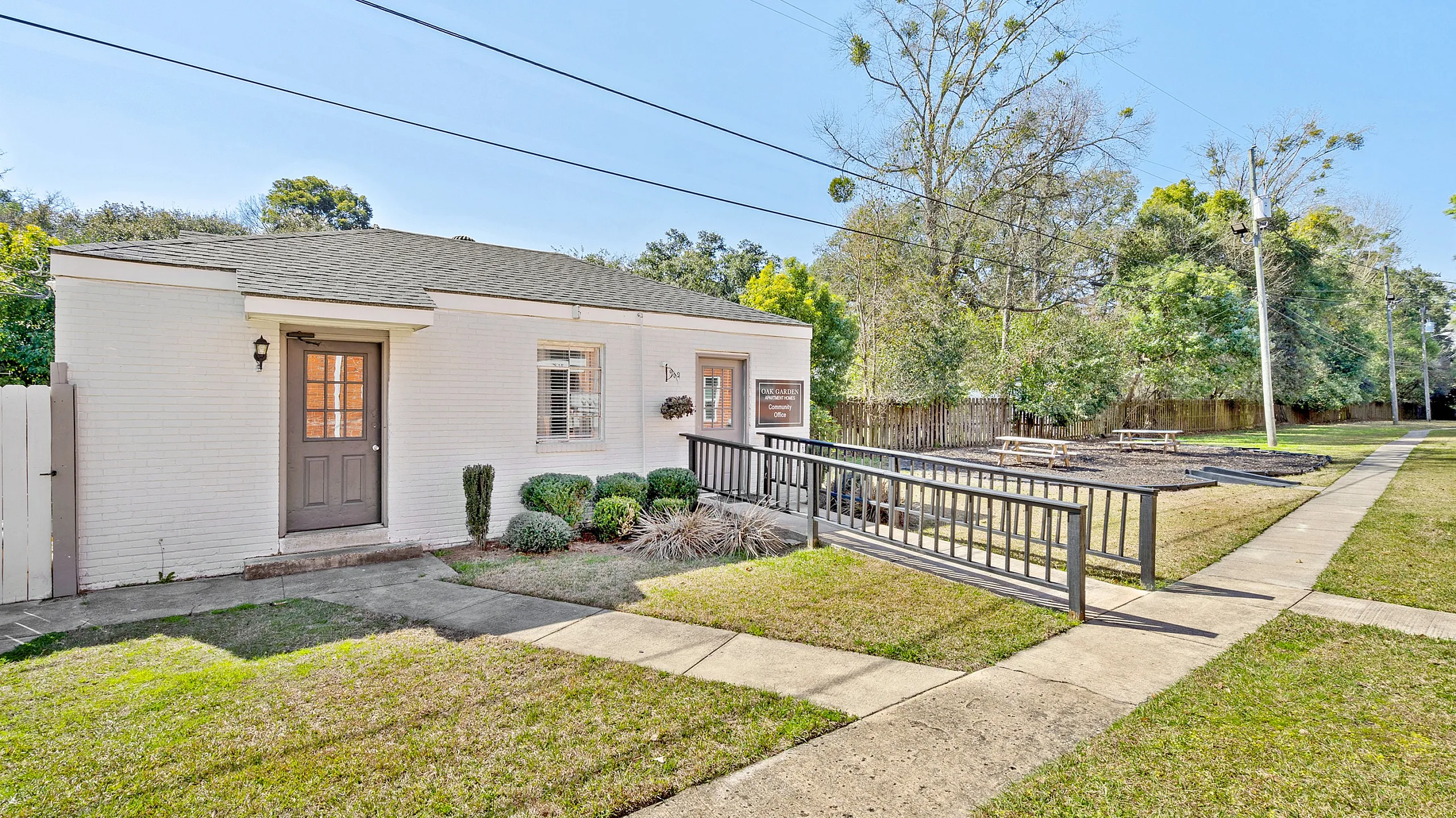 Charming white brick apartment with a welcoming front door, lush greenery, and modern accessibility ramp.