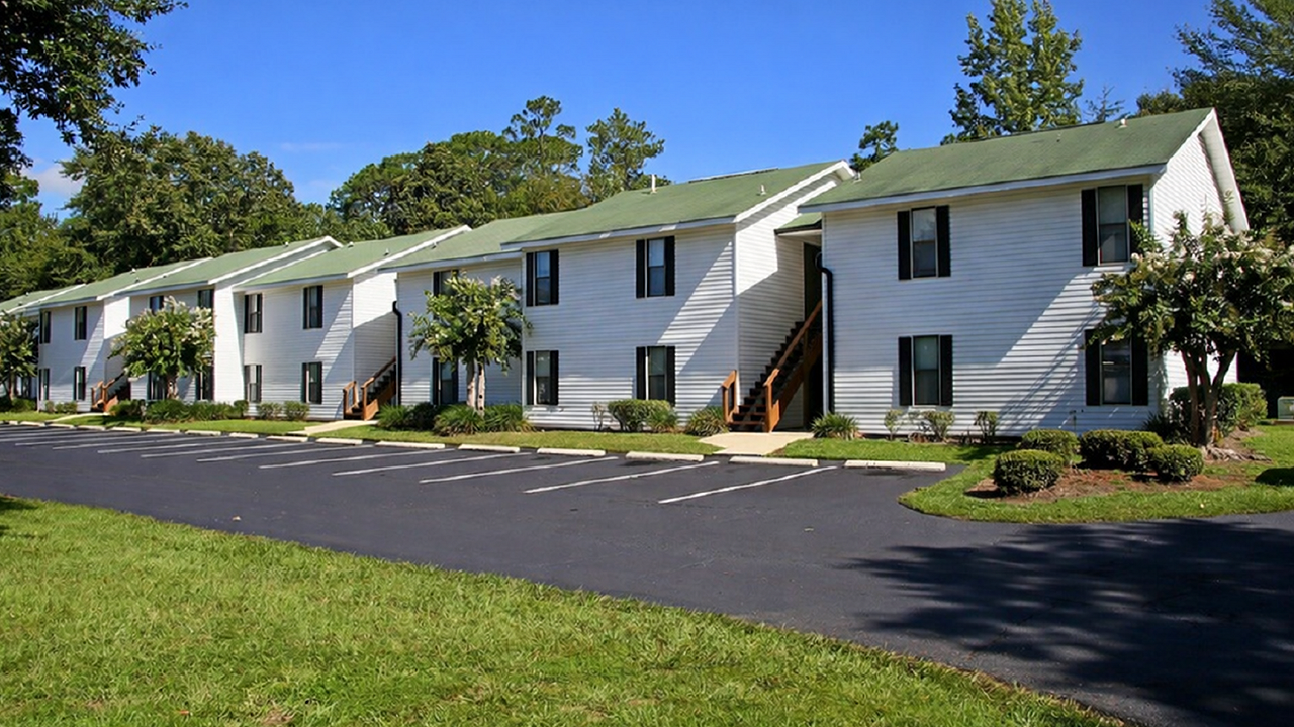 Charming apartment complex with white siding, lush landscaping, and ample parking, nestled in a serene green environment.