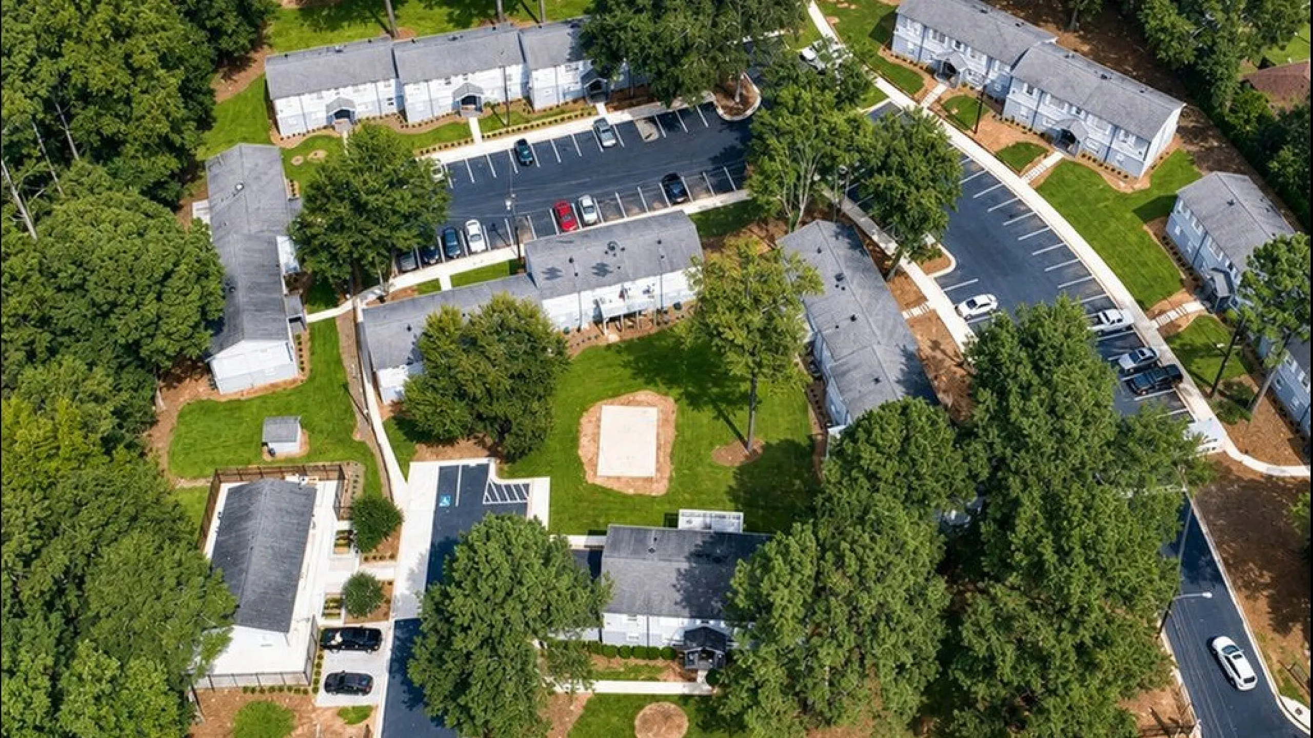 Aerial view of a charming apartment complex nestled in lush greenery, featuring well-maintained pathways, ample parking, and spacious communal areas.