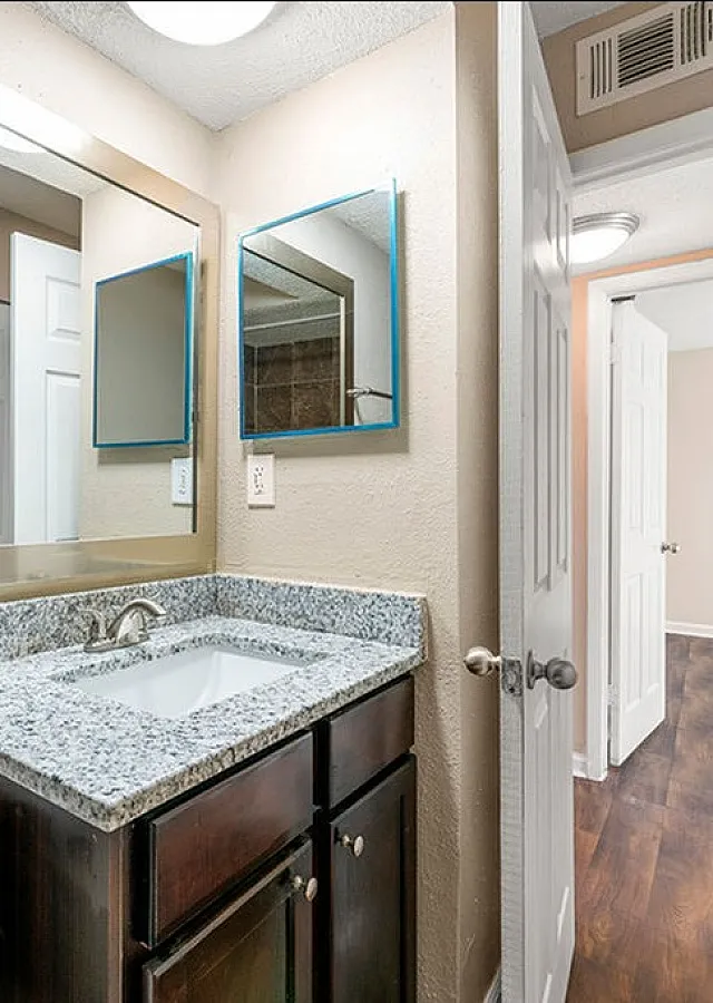 Bright and modern bathroom with granite countertop and elegant finishes, leading to a stylish wooden floored hallway.