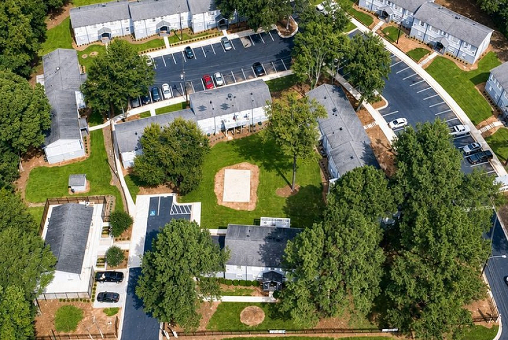 Charming aerial view of a serene apartment complex nestled in lush greenery, featuring tree-lined pathways and ample parking spaces.