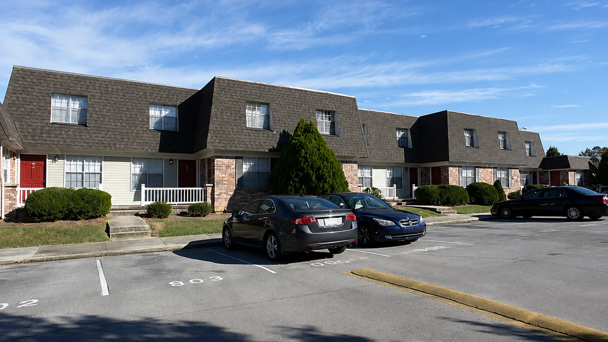 Charming townhouse complex with brick facades and cozy front porches, featuring ample parking and lush greenery under a clear blue sky.