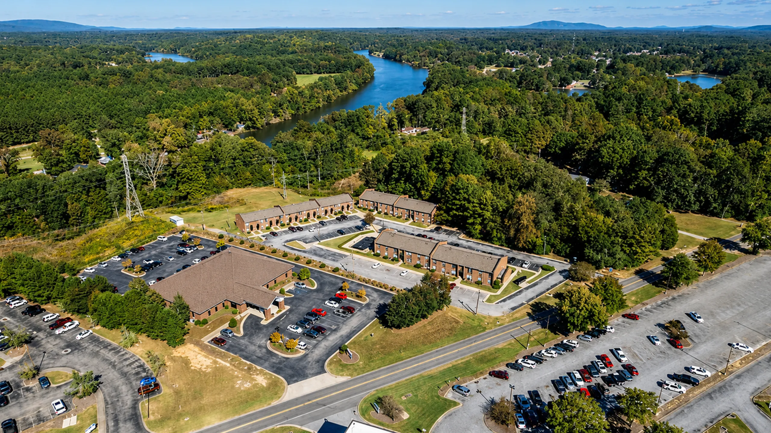 Scenic aerial view of a charming apartment complex nestled by a winding river, surrounded by lush greenery and ample parking.