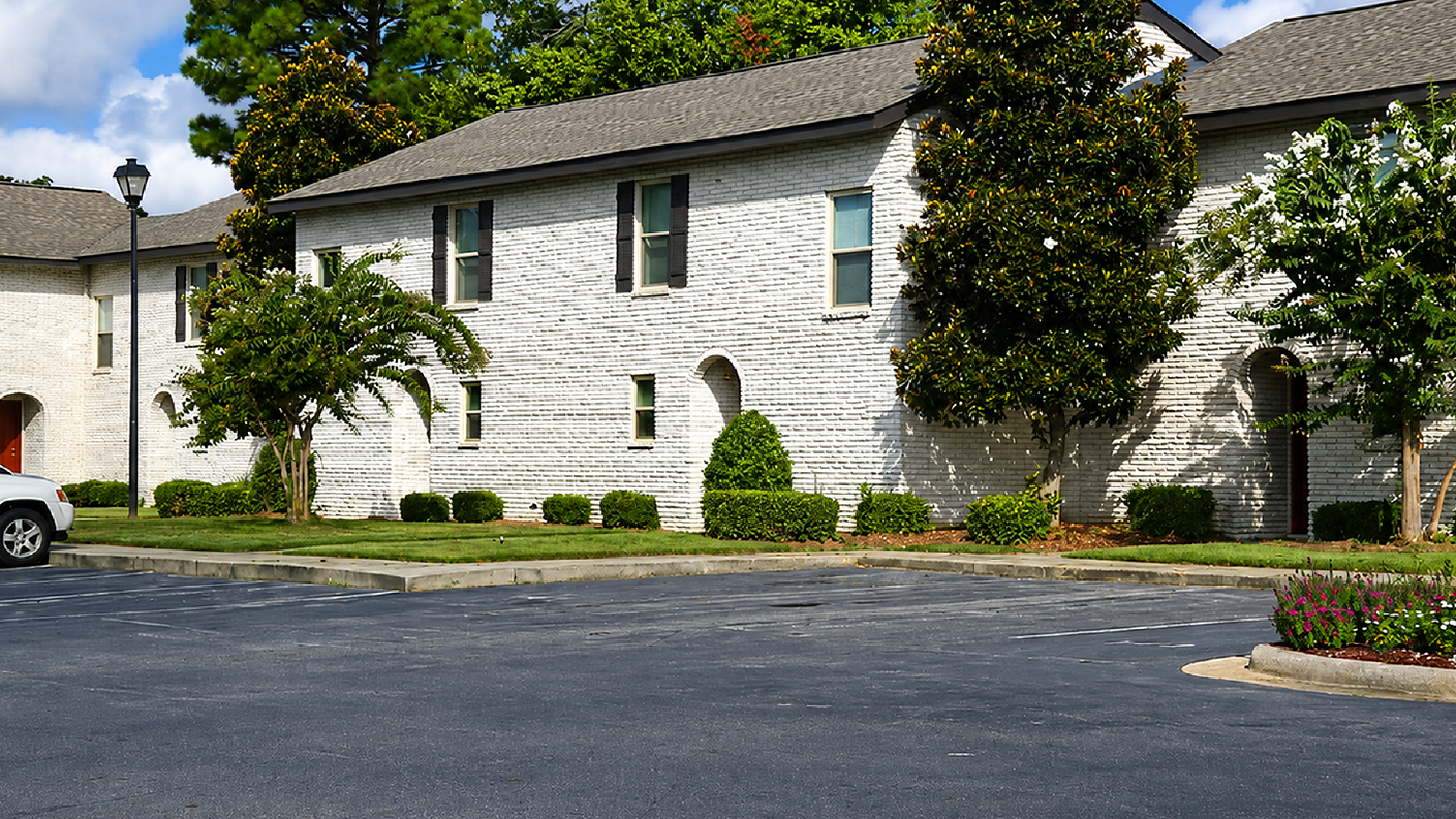 Charming two-story apartment building with classic white brick exterior, surrounded by lush greenery and vibrant landscaping, set against a blue sky.