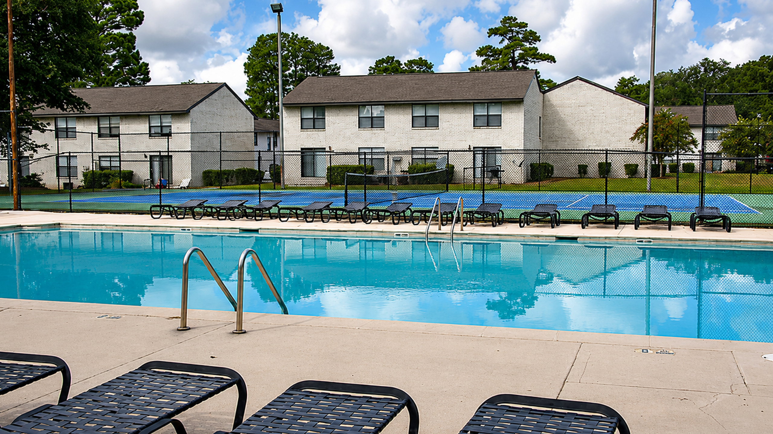 Inviting apartment complex with a sparkling swimming pool, surrounded by lounge chairs and tennis courts in a lush setting.