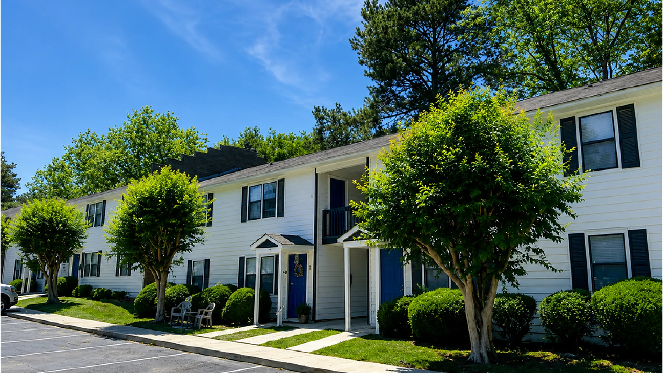 Charming apartment complex with vibrant greenery, featuring inviting blue doors and large windows under a brilliant blue sky.