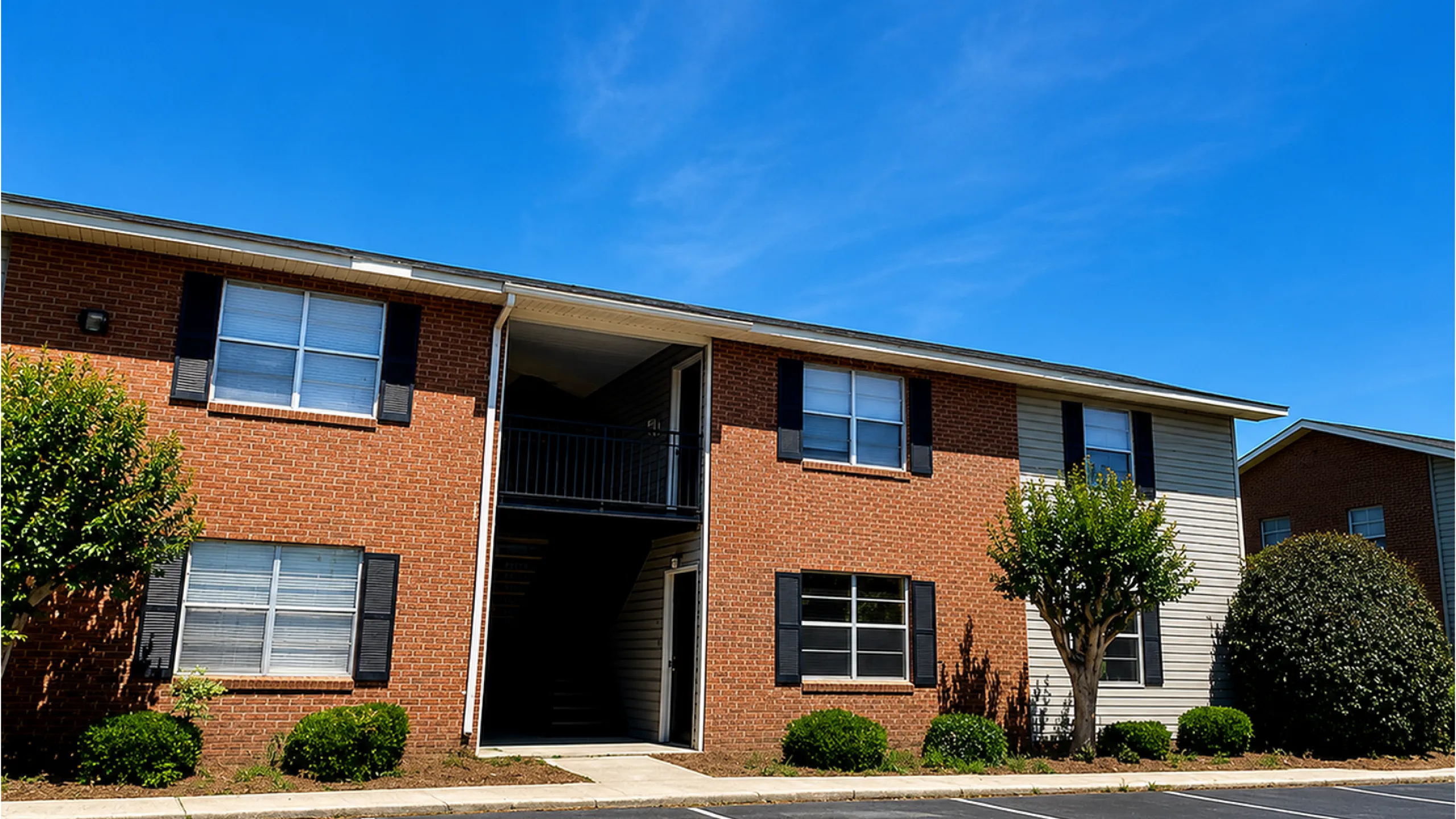 Charming brick apartment building with lush greenery and ample parking space, basking in bright sunlight.