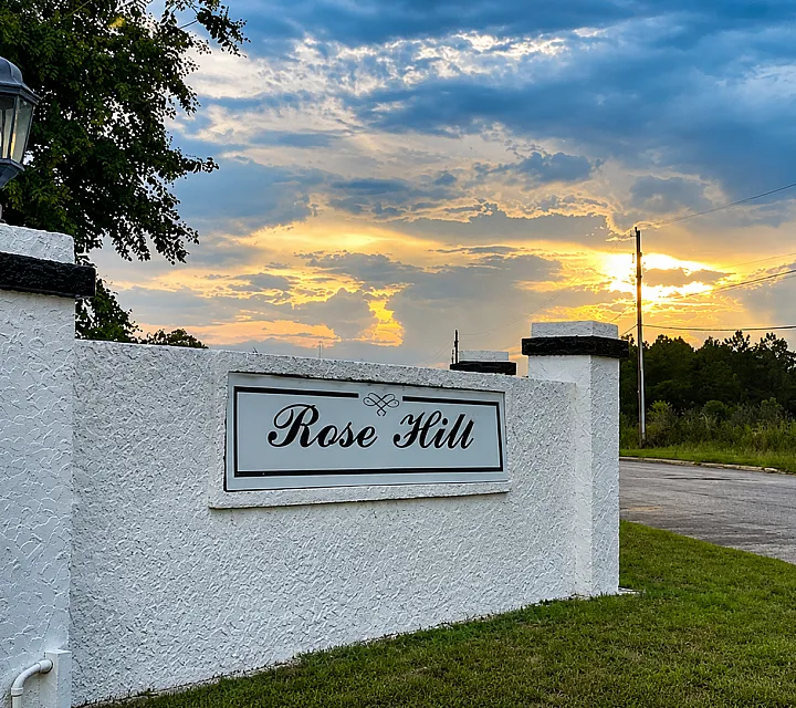 Charming entrance to Rose Hill with elegant sign, lush greenery, and a breathtaking sunset backdrop.
