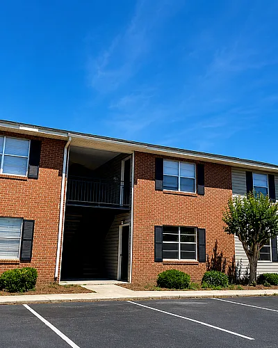 Charming brick apartment building with lush landscaping and ample parking, under a bright blue sky.