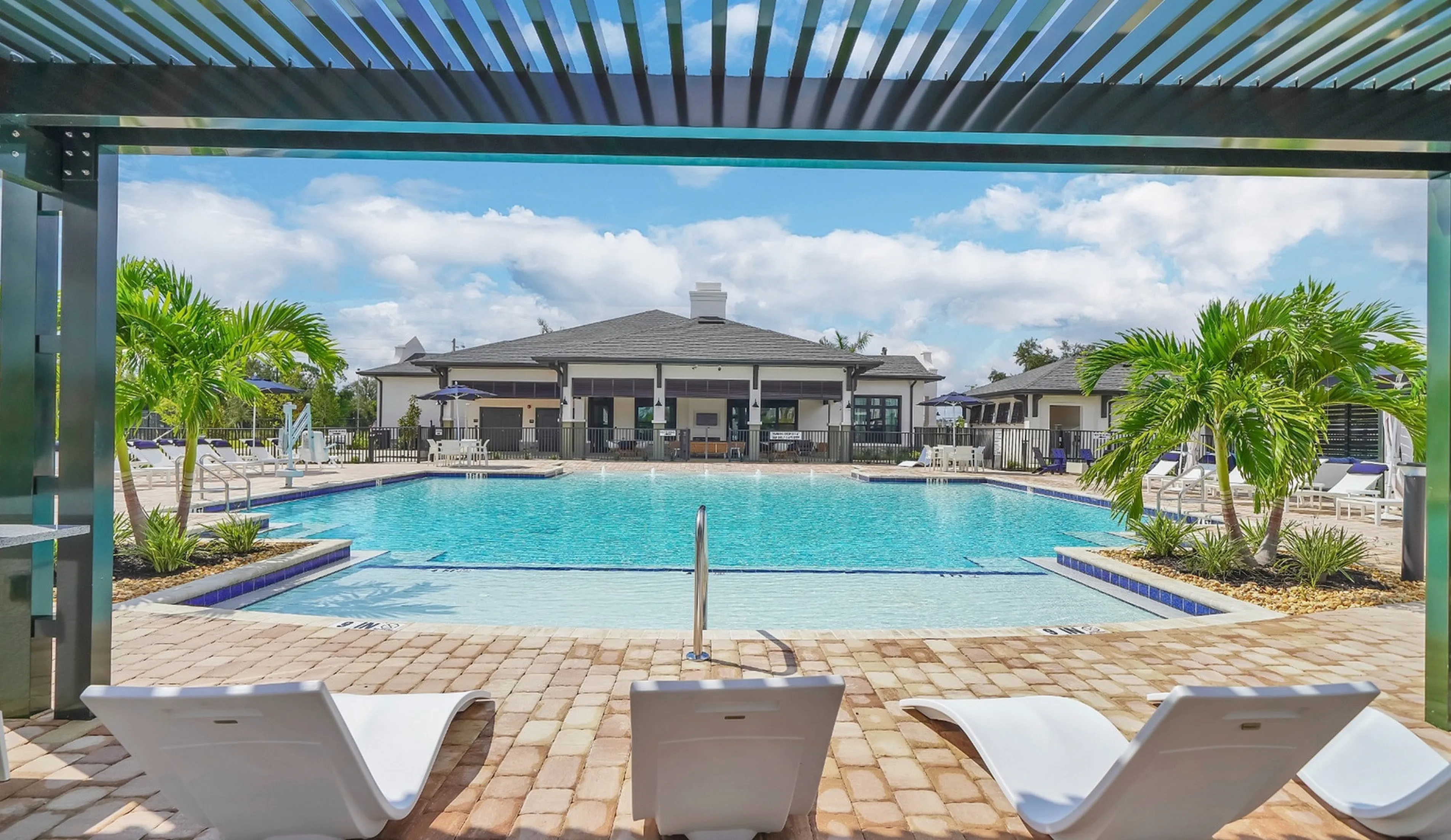 Luxury outdoor pool area with elegant lounge chairs, surrounded by lush palm trees and a modern clubhouse under blue skies.