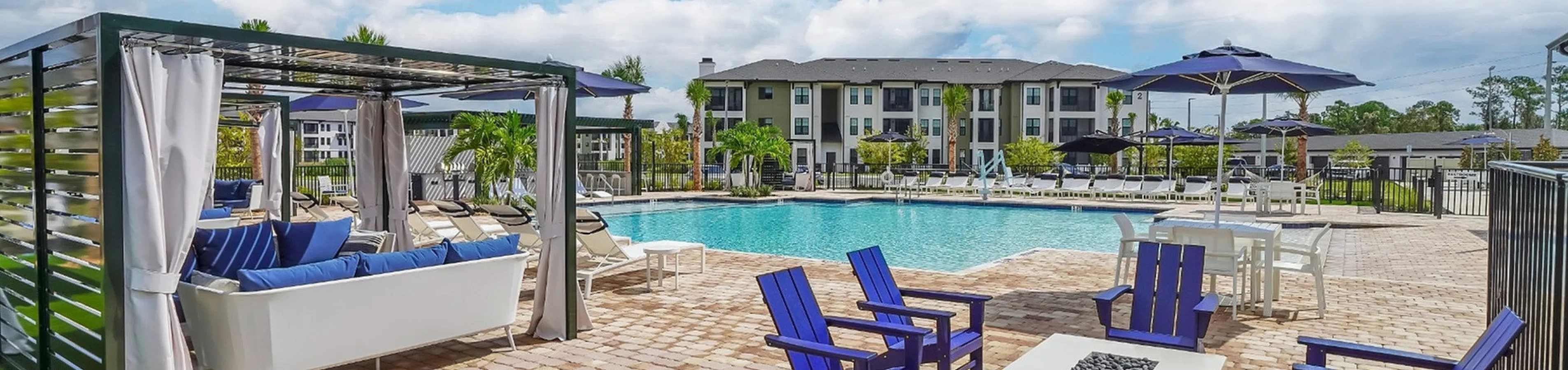 Luxurious outdoor pool area with elegant cabanas, vibrant blue lounge chairs, and a cozy fire pit, set against a backdrop of modern apartment buildings.