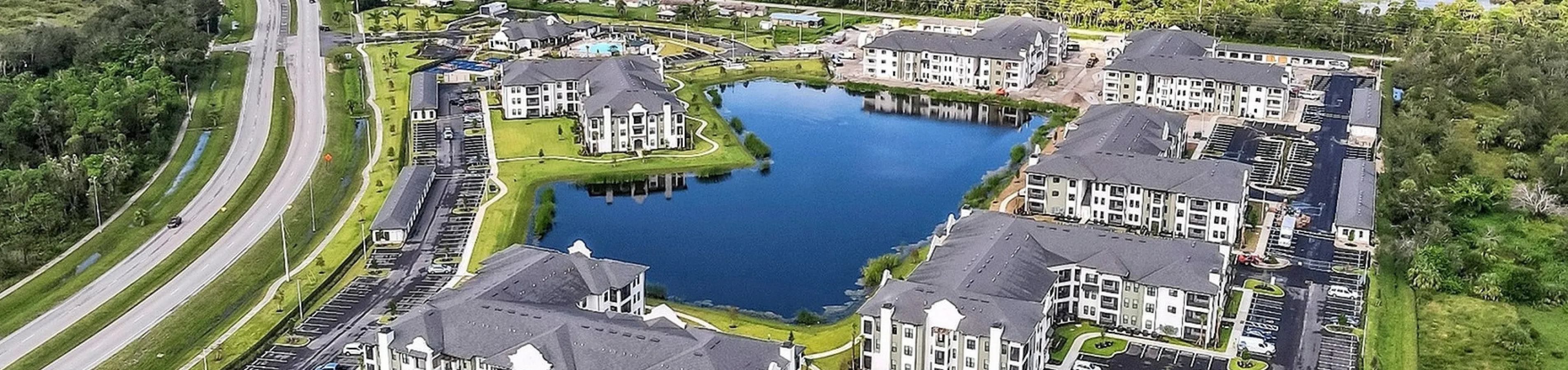 Aerial view of a luxurious apartment complex surrounded by lush greenery, featuring elegant buildings and a serene pond in the center.