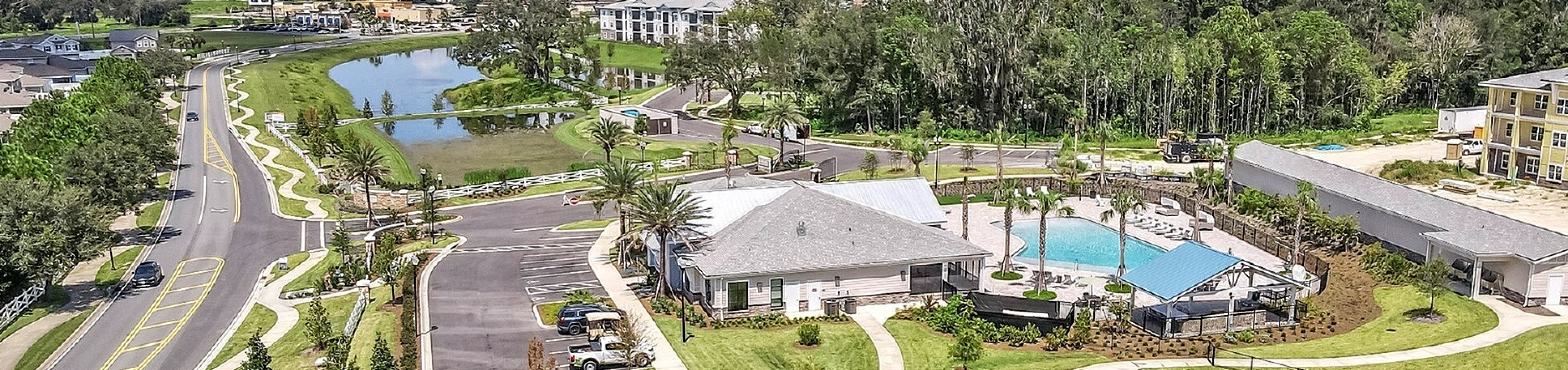 Beautiful aerial view of a luxurious apartment complex featuring a sparkling pool, lush green tennis courts, and scenic landscaping surrounded by serene water views.
