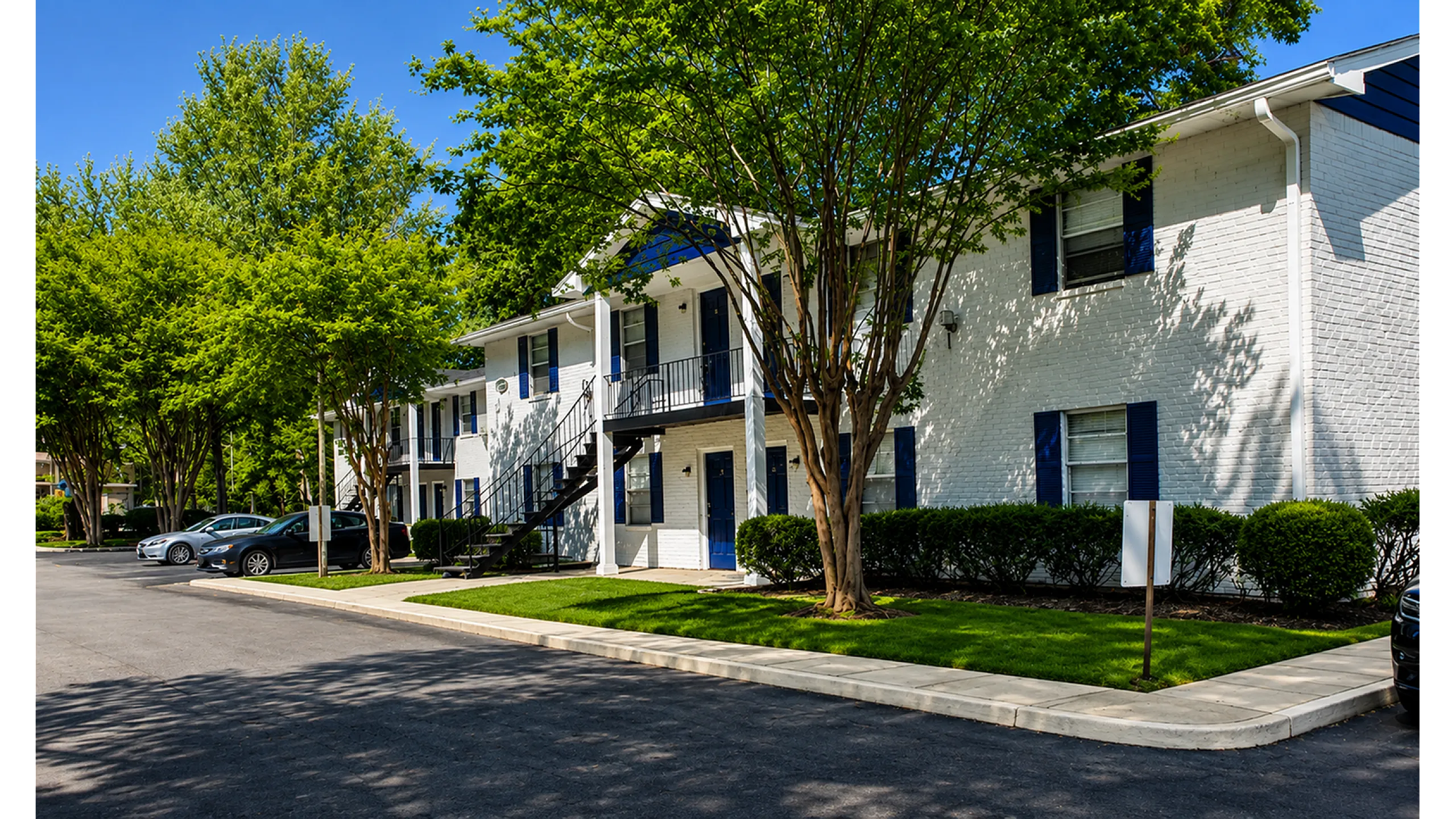 Charming two-story apartment building with blue shutters, nestled in lush green surroundings and ample parking space.