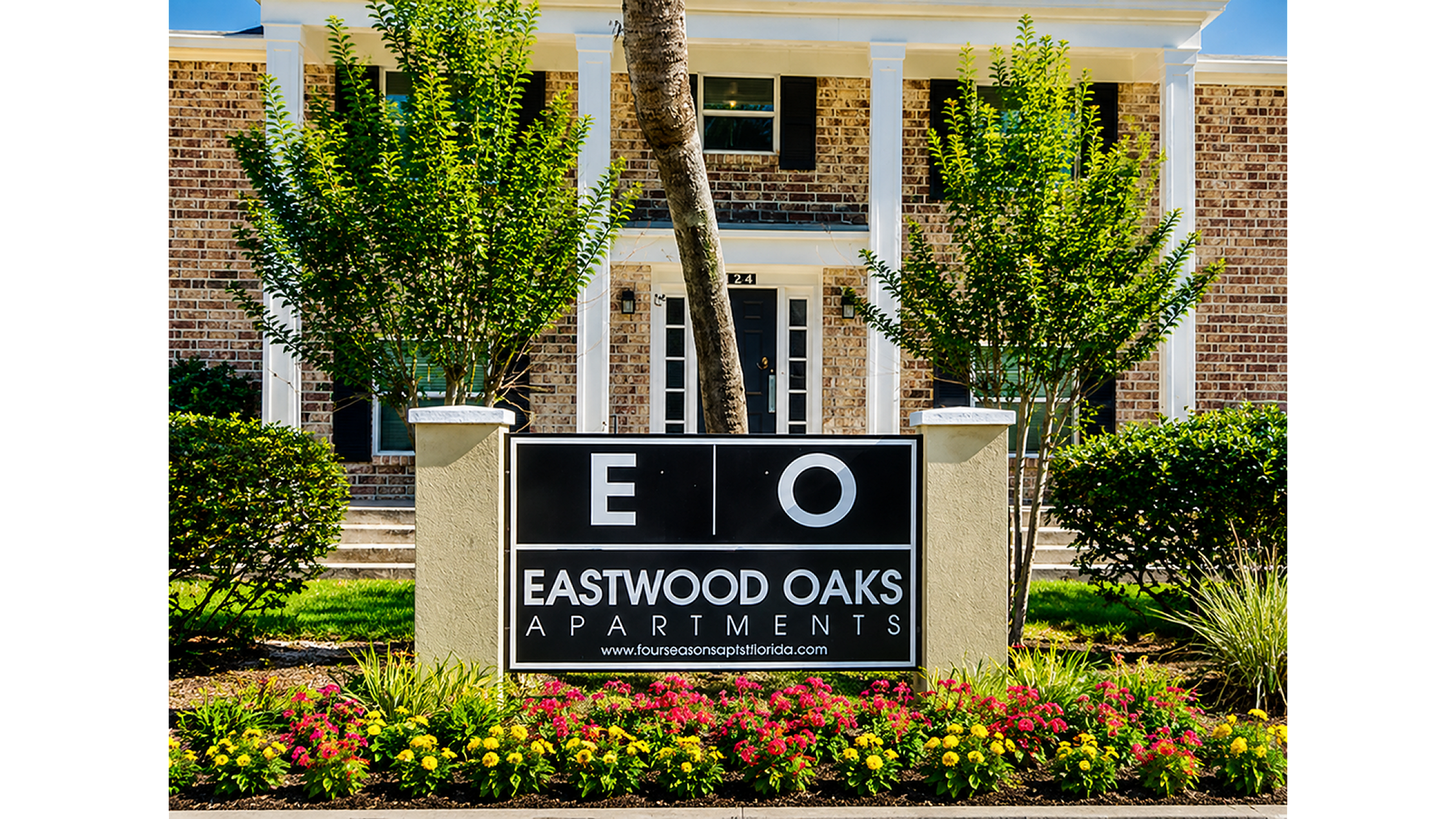 Charming entrance of Eastwood Oaks Apartments framed by lush greenery and colorful flowers, featuring elegant columns and a stylish brick facade.