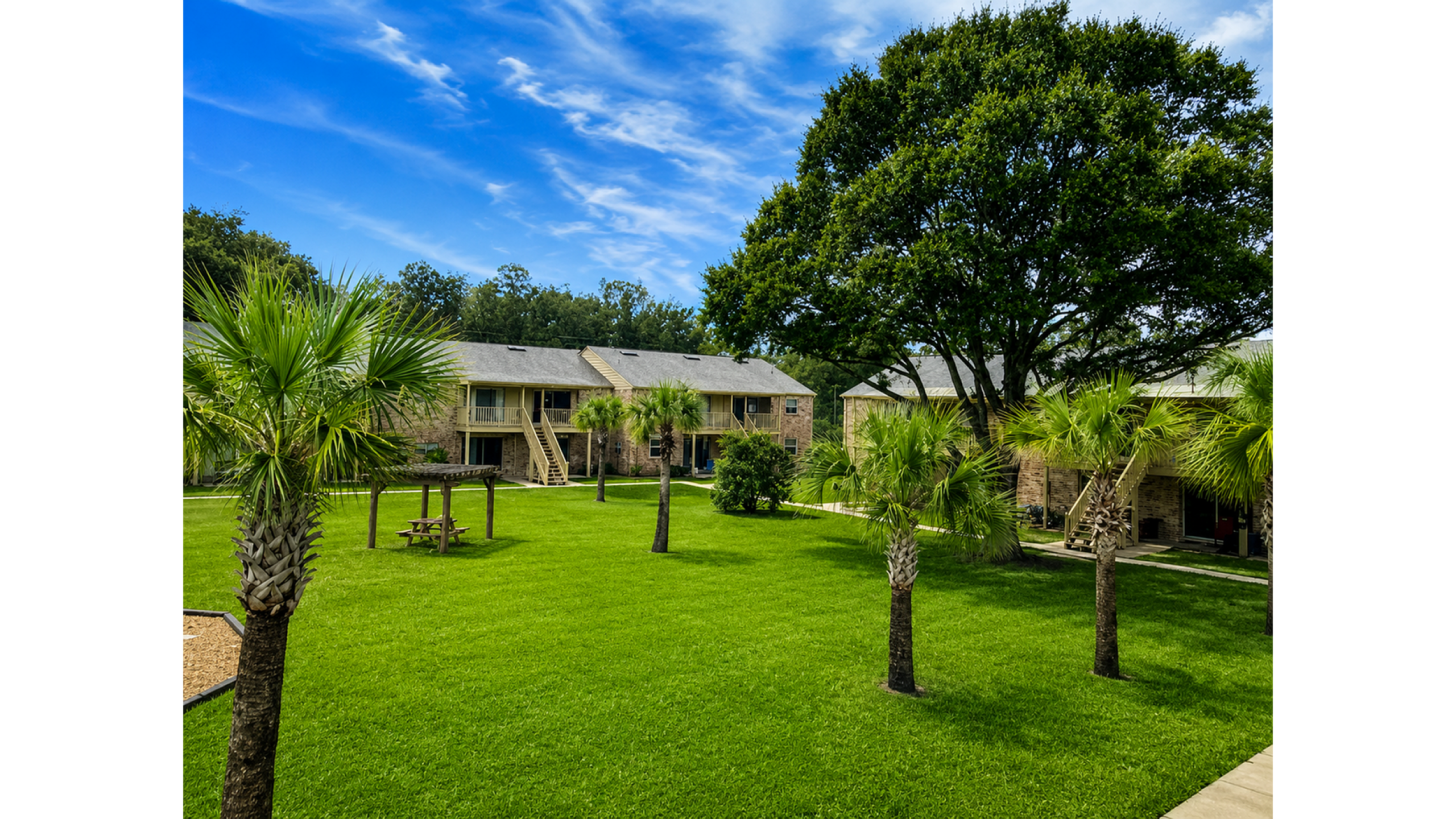 Charming apartment complex with lush green lawns, mature trees, and inviting outdoor seating areas under a vibrant blue sky.