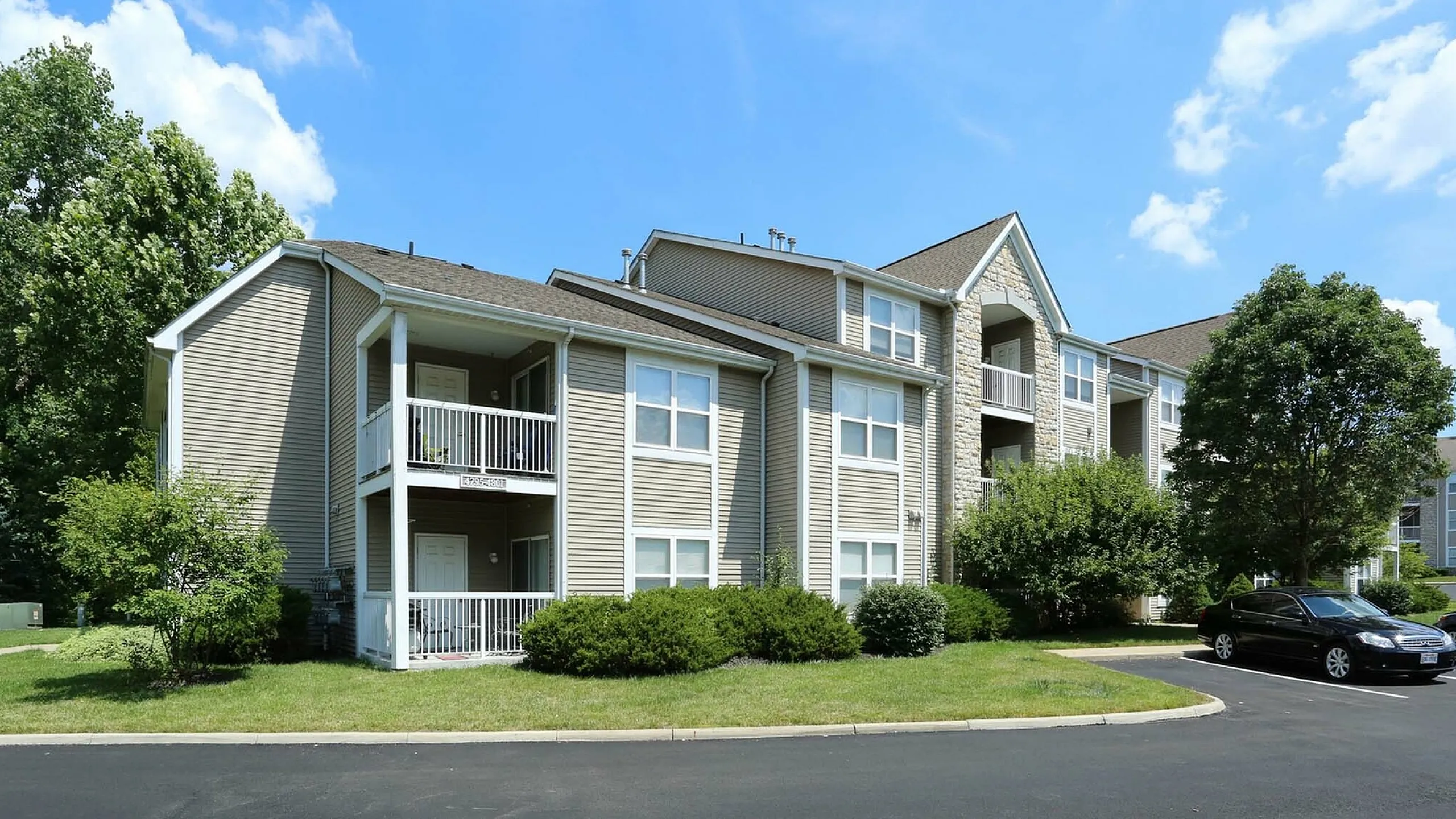 Charming apartment complex with modern facade, lush greenery, and convenient parking space under a bright blue sky.