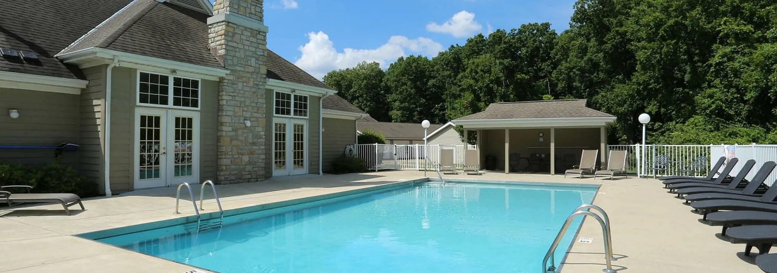 Inviting outdoor pool area with relaxing lounge chairs, surrounded by lush greenery and a charming stone clubhouse.