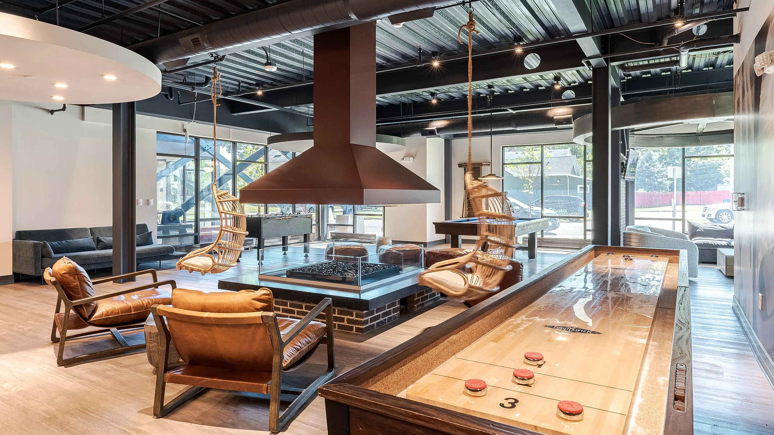 Chic modern lounge with leather seating, cozy fireplace, stylish swing chairs, shuffleboard table, and ample sunlight.