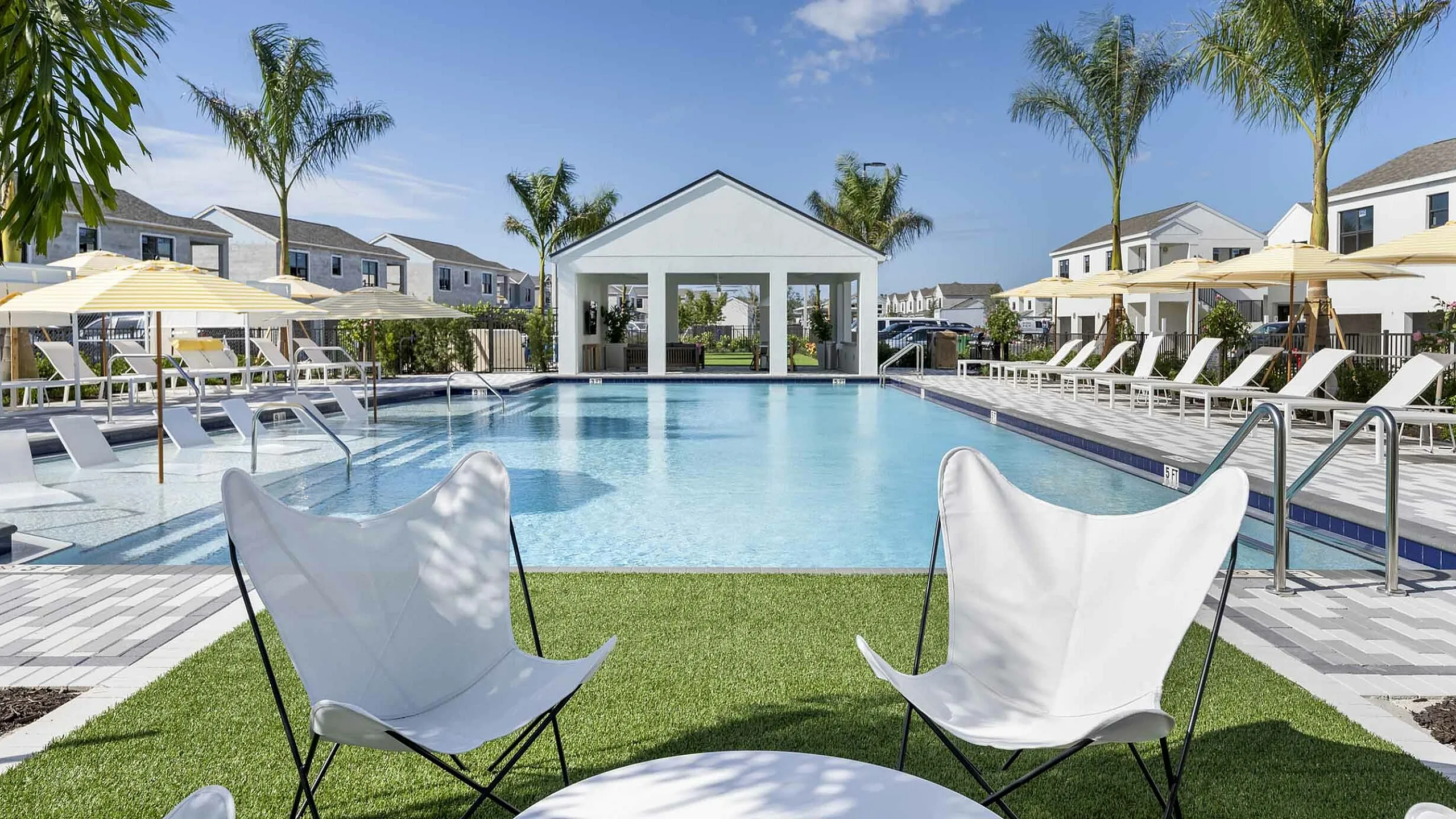 Luxurious community pool area with palm trees, chic lounge chairs, and modern architecture under a blue sky.