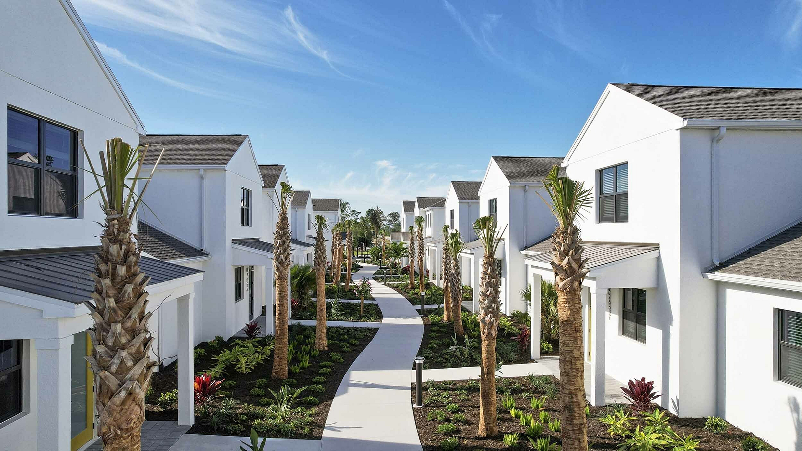 Stunning suburban neighborhood with modern white townhouses, lush landscaping, and palm-lined walkways under a clear blue sky.