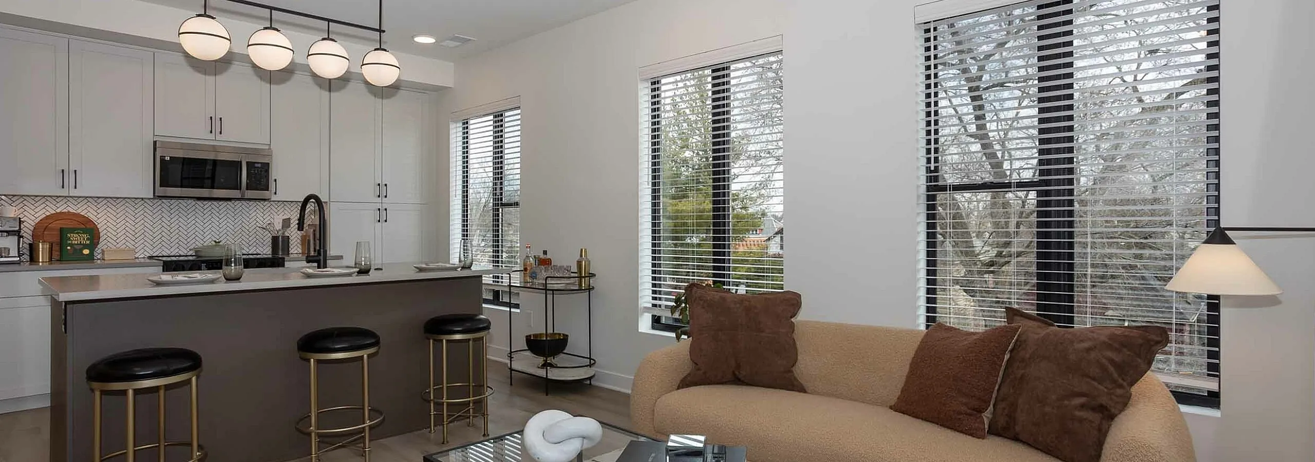 Modern open-concept living room and kitchen featuring chic pendant lighting, elegant bar stools, and large windows offering natural light.