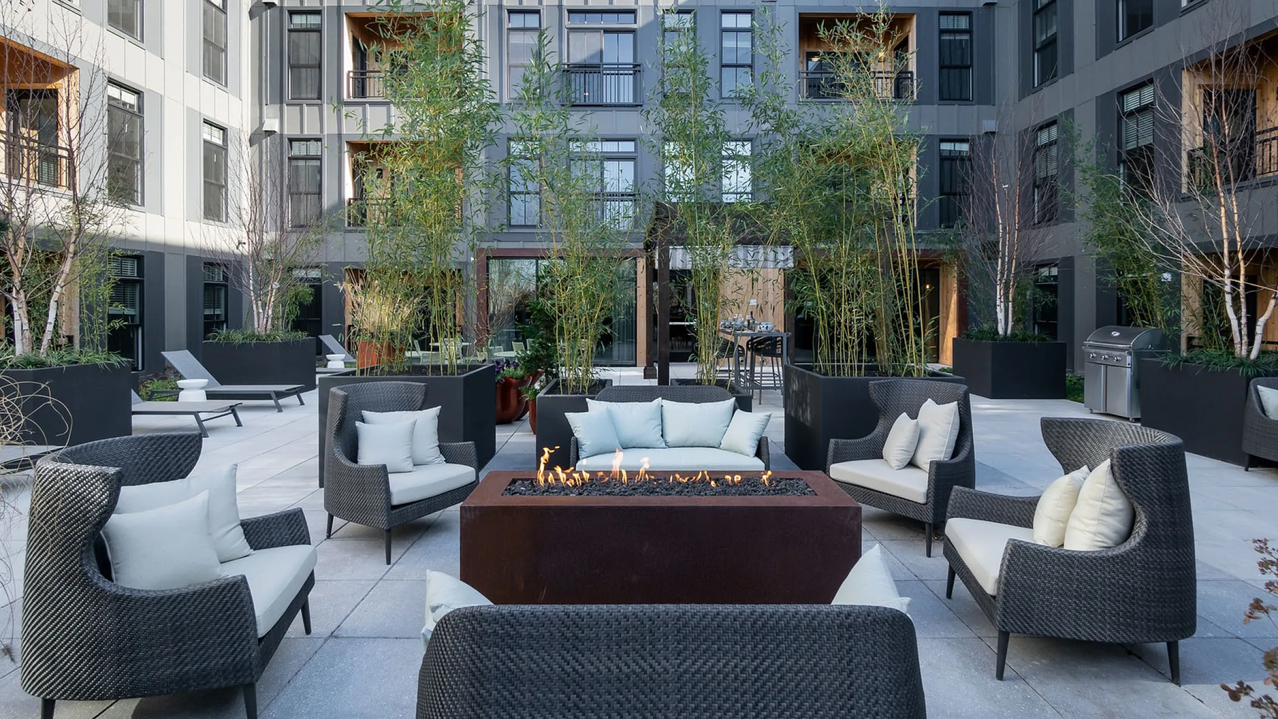 Elegant outdoor courtyard featuring modern wicker seating around a central fire pit, surrounded by lush greenery in a stylish urban apartment complex.