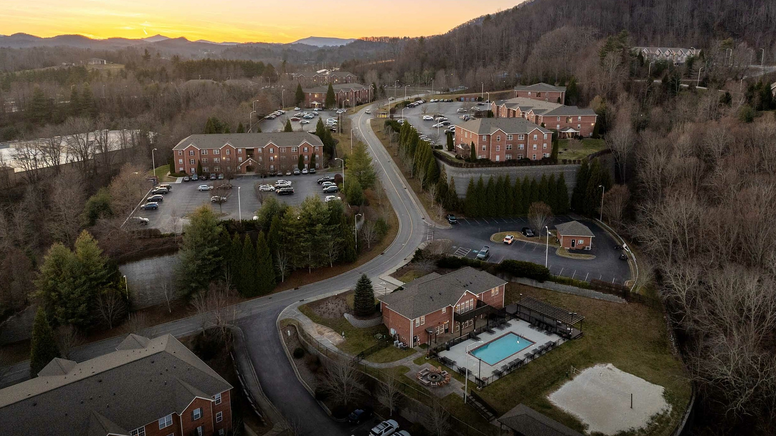 Scenic aerial view of a charming apartment community nestled in lush greenery, featuring well-maintained buildings, ample parking, and a refreshing swimming pool, all set against a breathtaking mountain backdrop at sunset.
