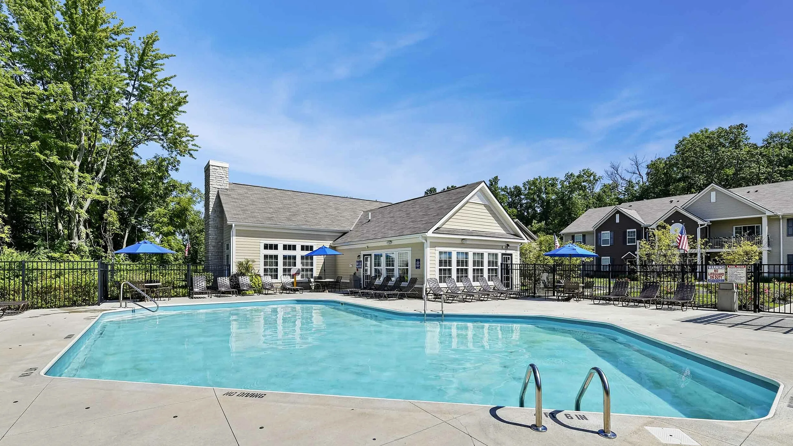 Inviting outdoor pool area with modern deck chairs and charming clubhouse, surrounded by lush greenery under a clear blue sky.