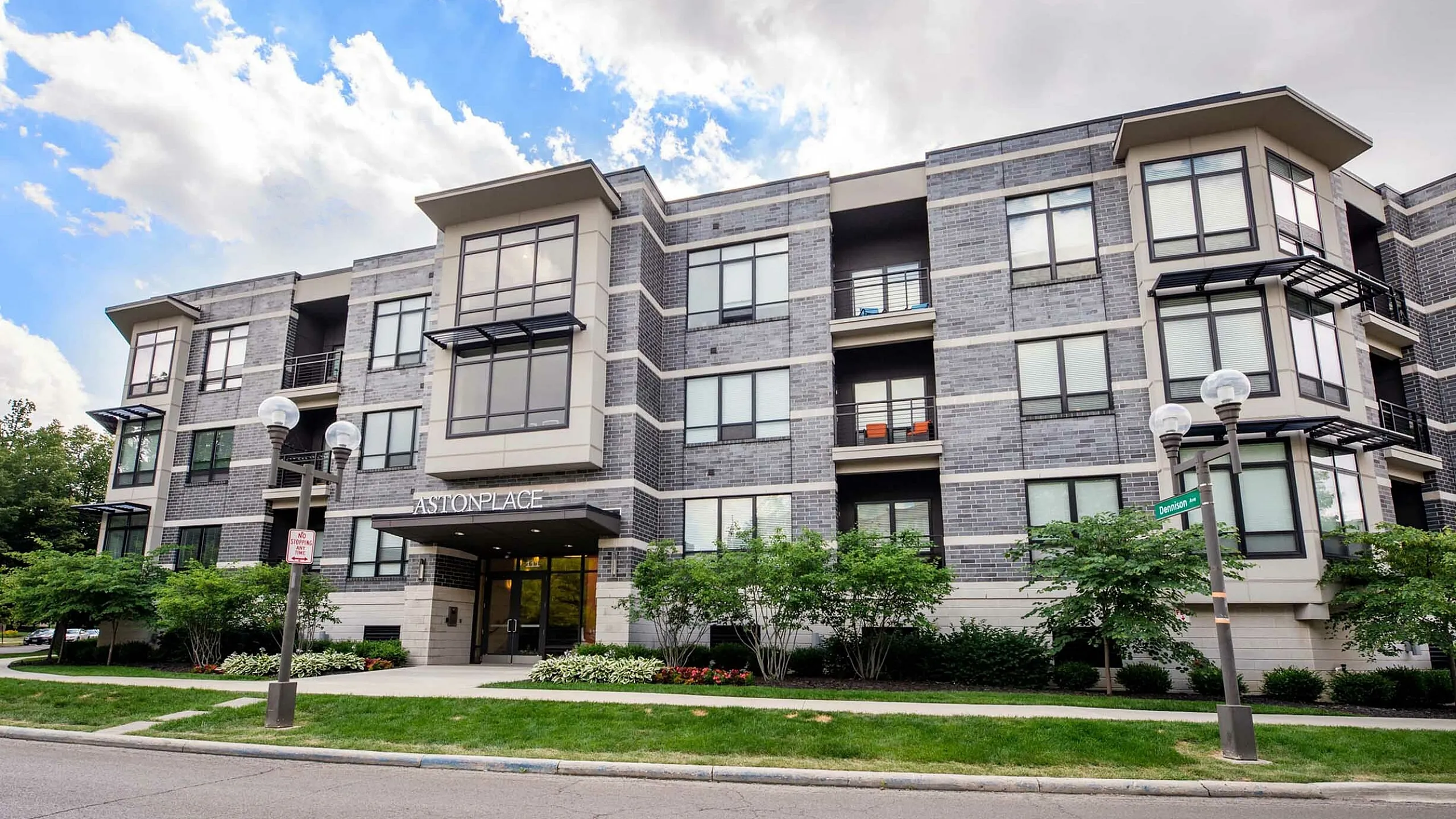 Modern apartment building with sleek design, large windows, and lush landscaping at Aston Place under a bright blue sky.