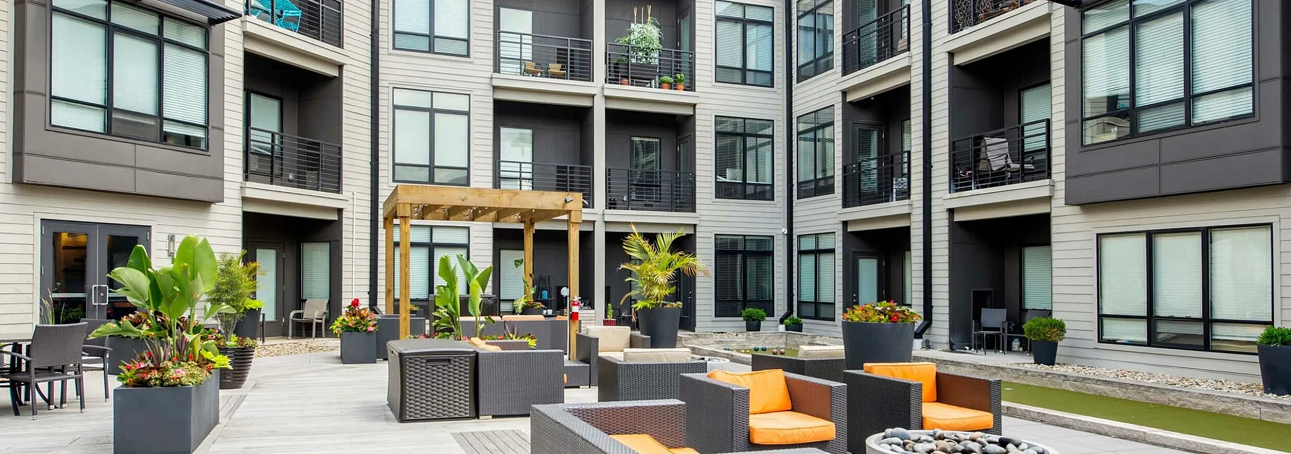 Modern apartment courtyard with stylish outdoor seating, lush greenery, and chic architecture under a bright sky.