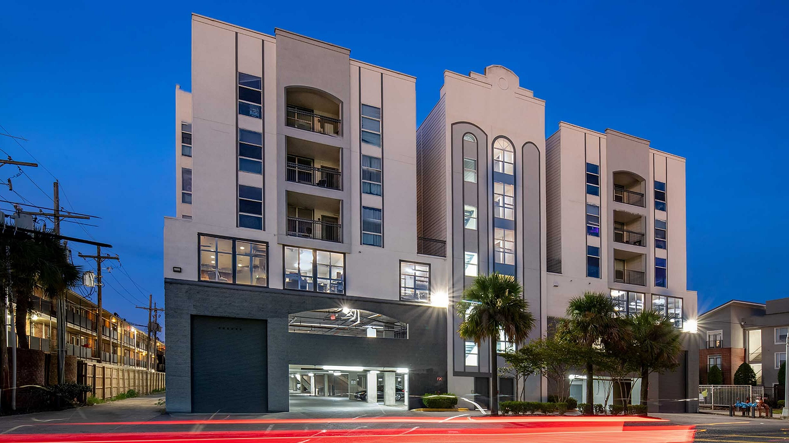 Modern urban apartment building with elegant architectural design, featuring well-lit windows, surrounded by palm trees and city nightlife with dynamic light trails.