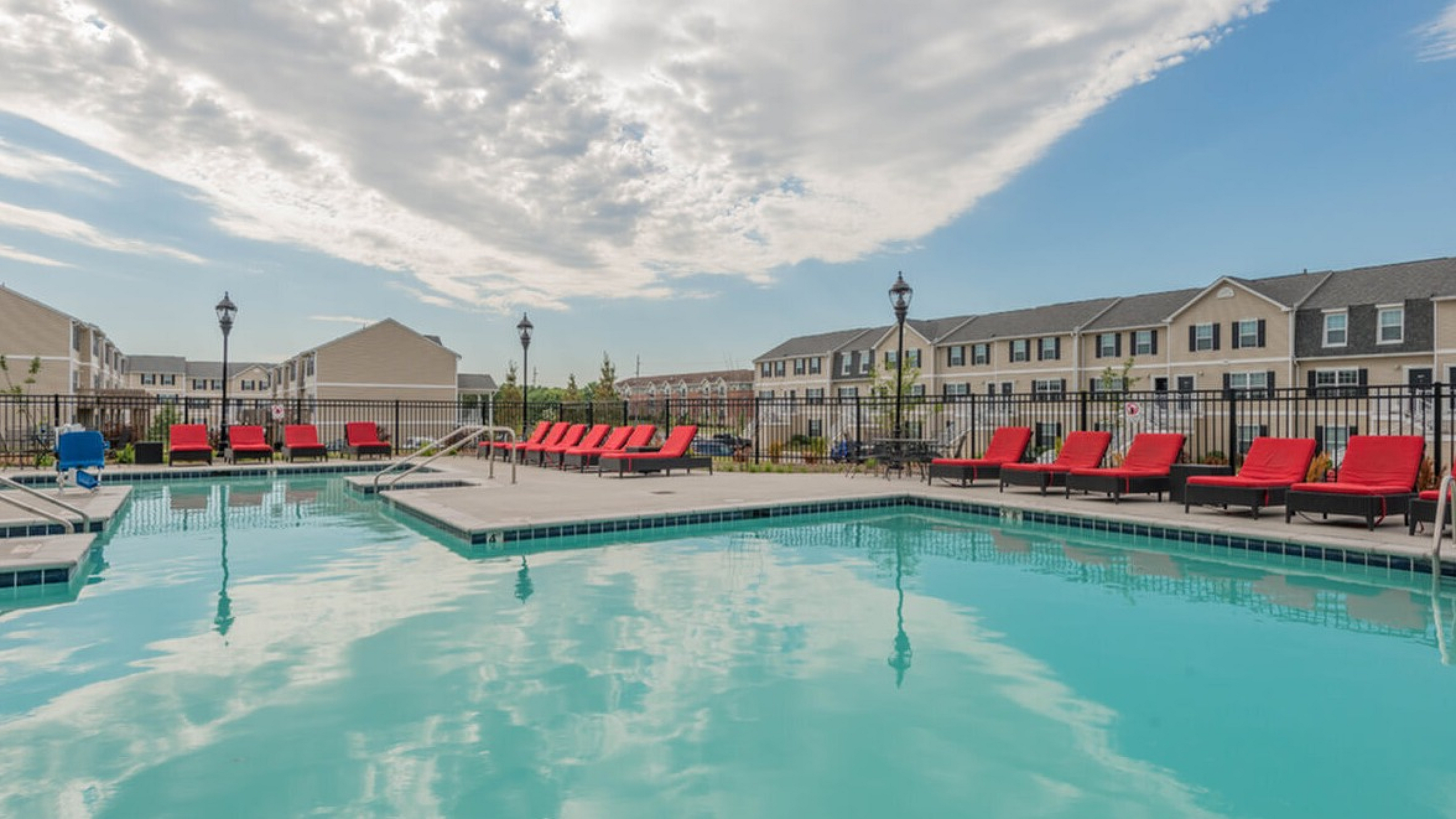Inviting apartment complex with a sparkling swimming pool, surrounded by red lounge chairs and sunlit townhouses in the background.