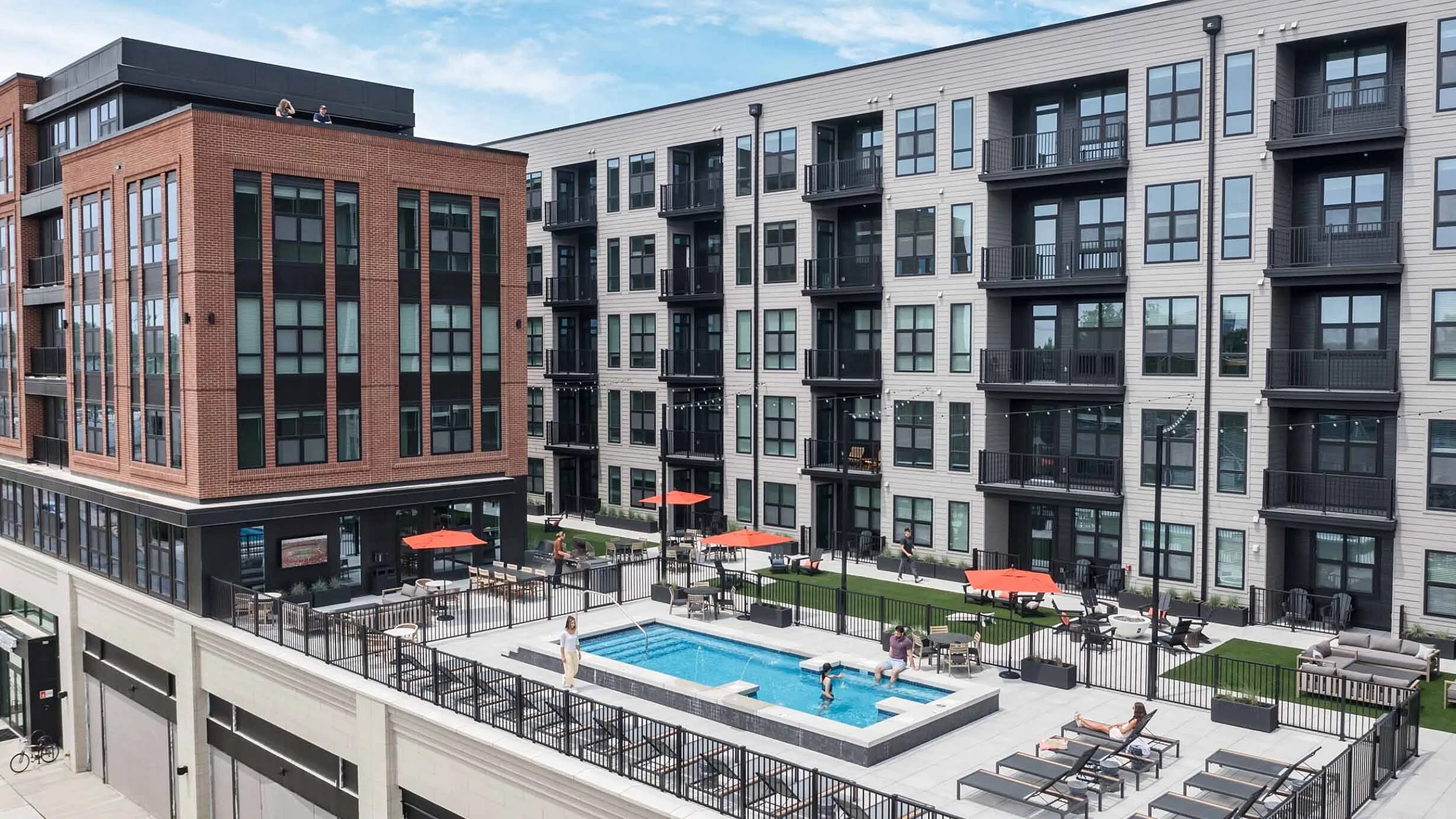 Modern apartment complex with stylish brick and siding exterior, featuring a luxurious rooftop pool and lounge area adorned with red umbrellas.