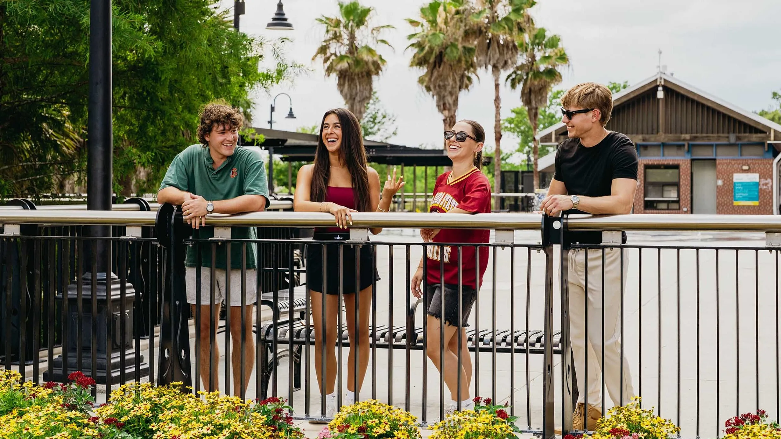 Group of friends laughing and enjoying a sunny day by a vibrant flower garden and modern lampposts.
