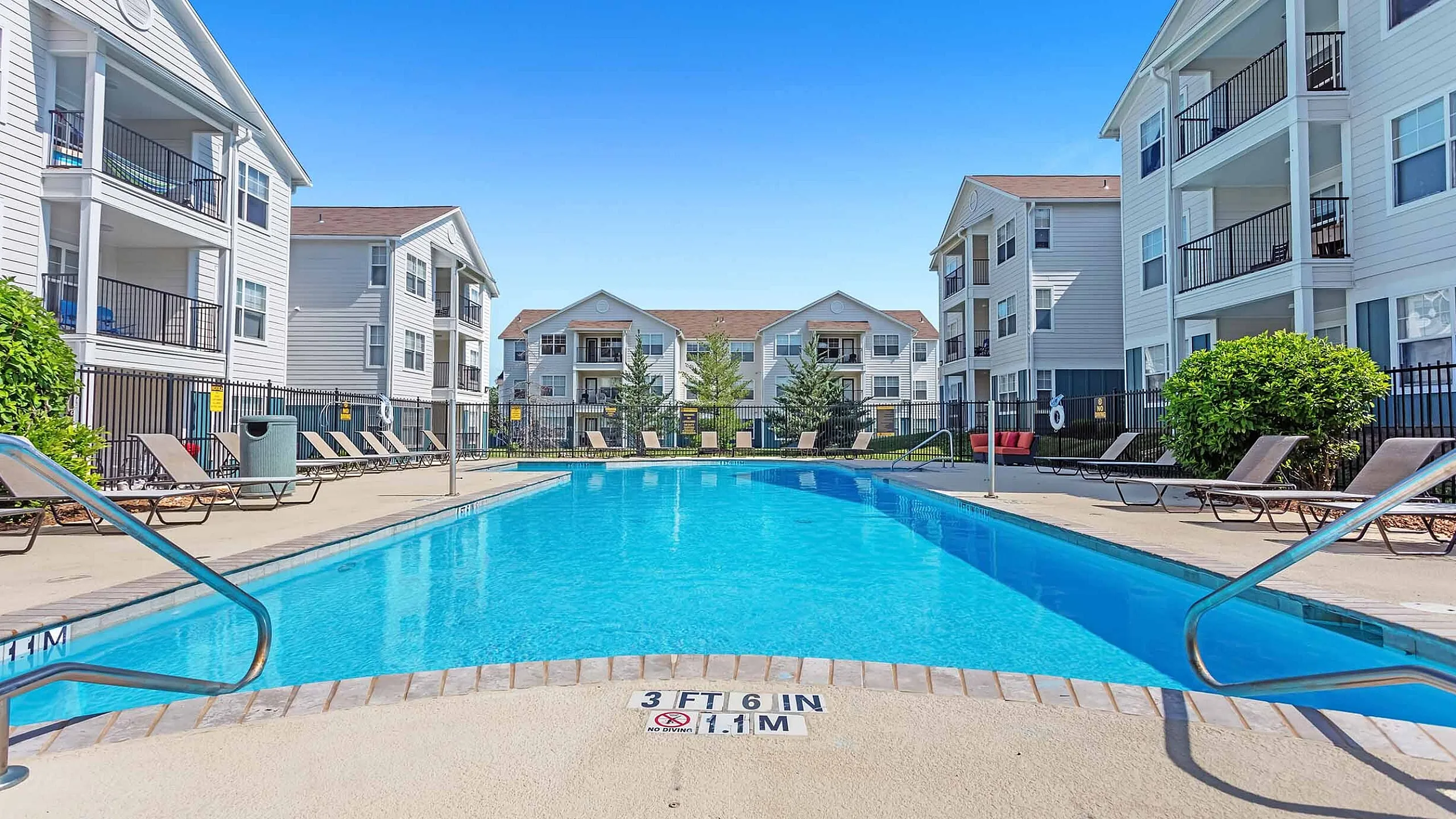 Luxury apartment complex with sparkling swimming pool, surrounded by modern three-story buildings and cozy lounge chairs.