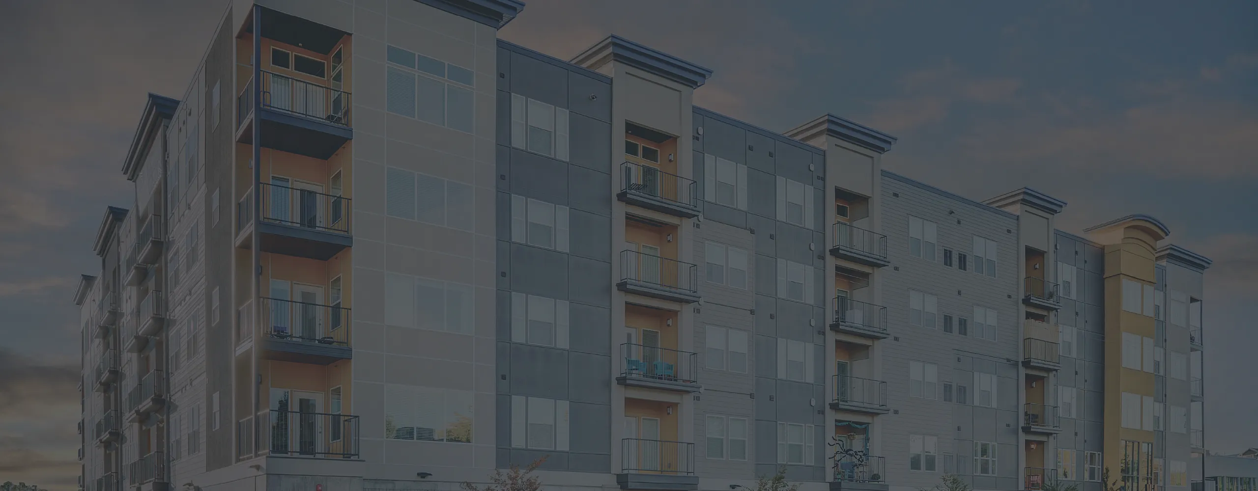 Modern multi-story apartment building with sleek architecture and vibrant balconies against a colorful evening sky.