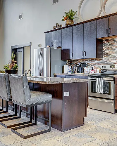 Elegant modern kitchen with sleek dark cabinetry, a stylish backsplash, and a spacious island with leather bar stools, adjacent to a cozy living area with large windows.